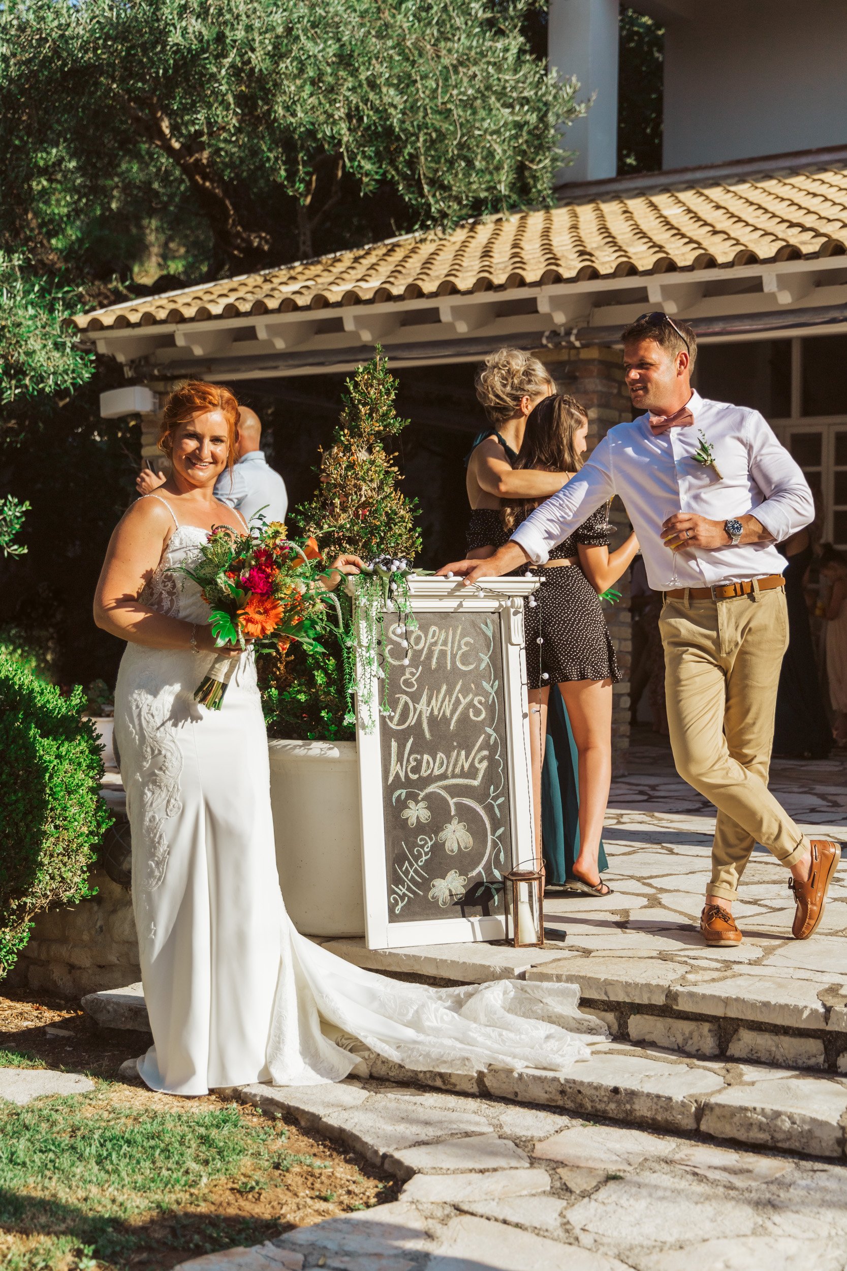 Bride holding a bouquet smiling near a chalkboard sign at an outdoor wedding reception with guests chatting in the background.