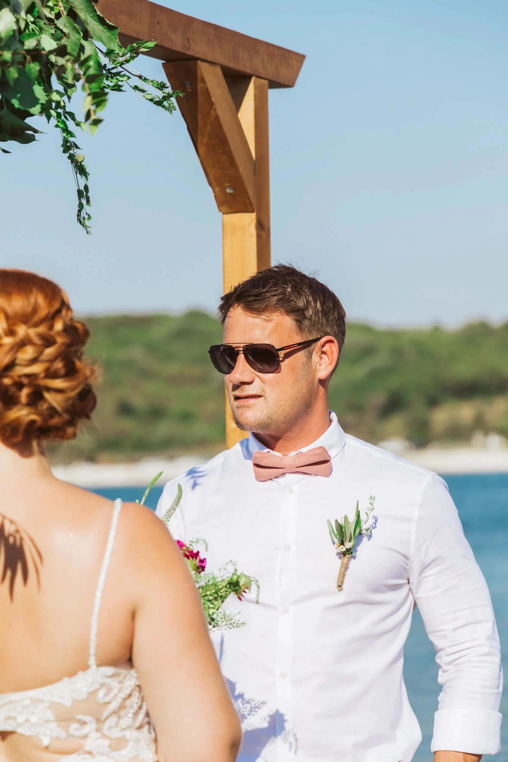 A man wearing sunglasses, a white shirt, and a pink bow tie, standing outdoors near water with a woman who has red hair styled in an updo wearing a lace dress. They appear to be participating in a wedding ceremony.