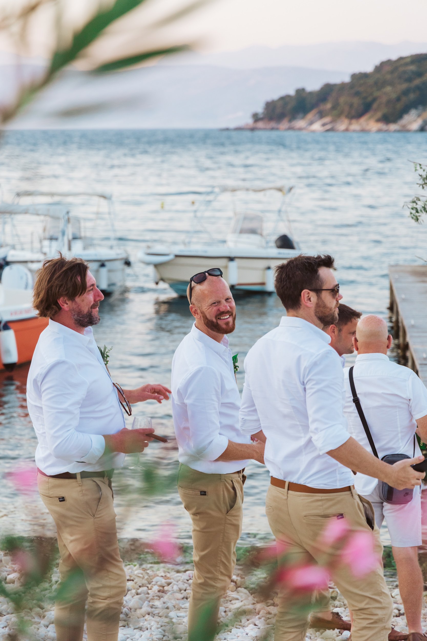 Group of men in white shirts and khaki pants standing near a dock by the water, boats in the background, during sunset or late afternoon.