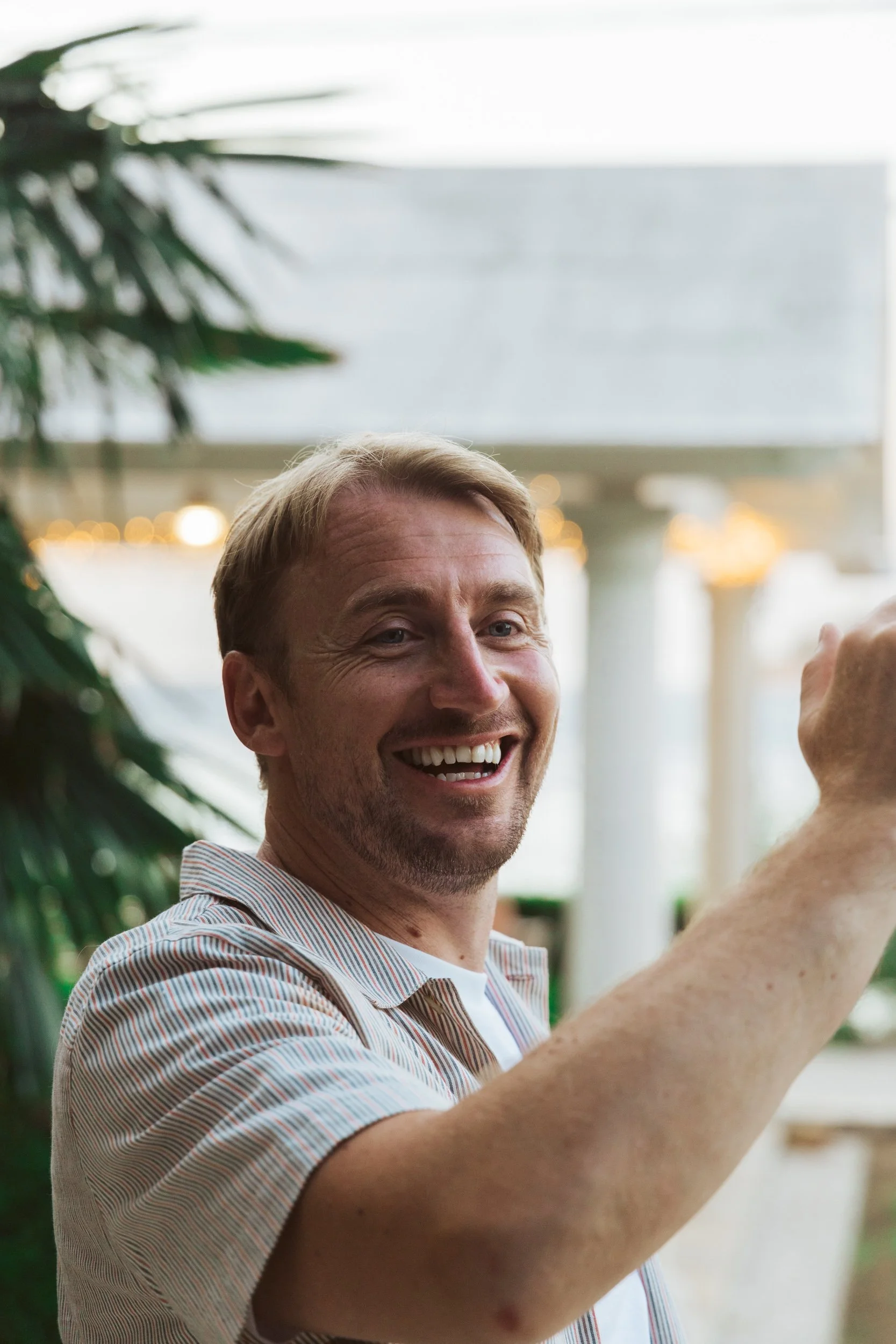 A man smiling and waving outdoors with palm leaves and white columns in the background.