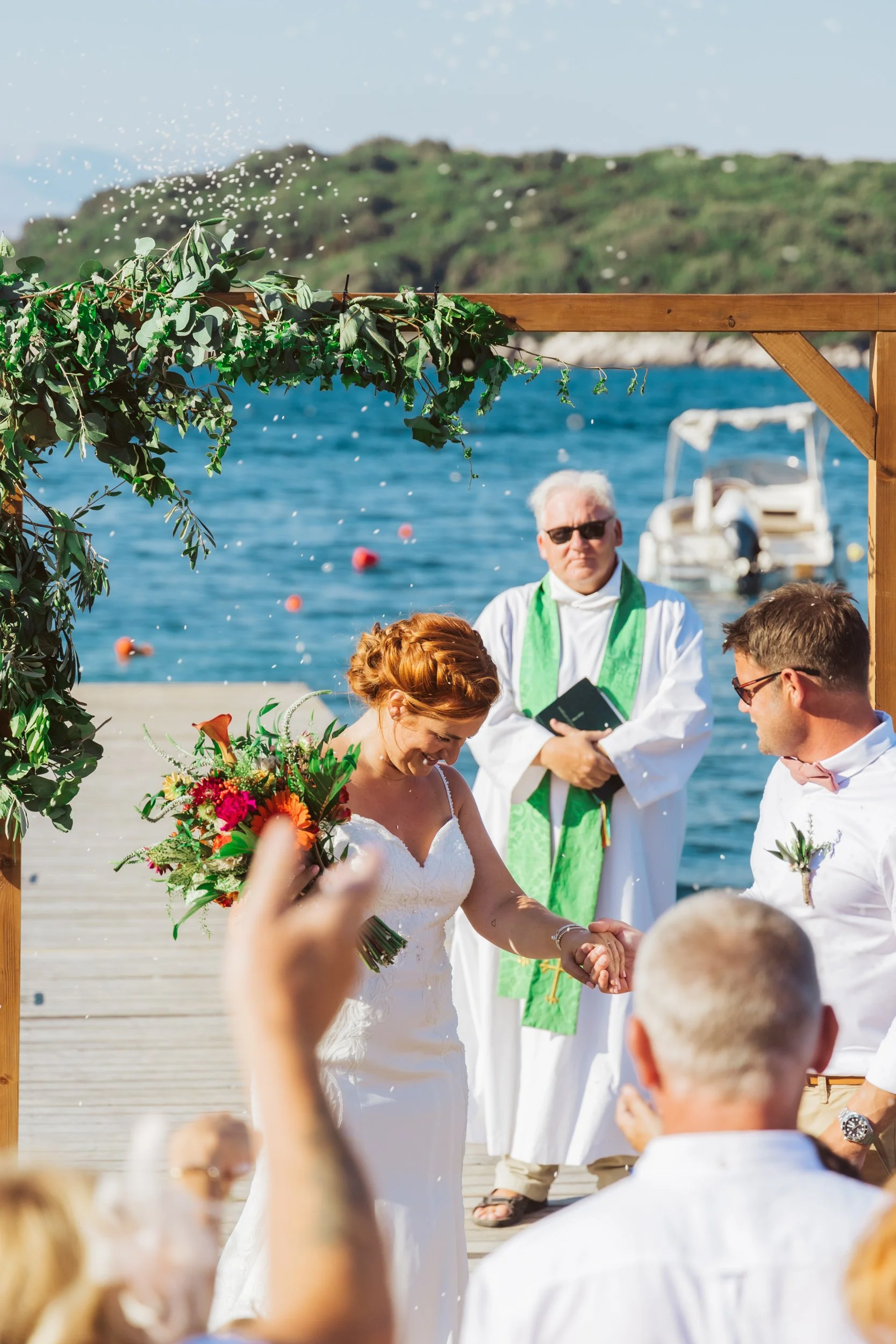 A couple getting married outdoors at a seaside wedding, with a priest officiating, guests celebrating, and boats on the water in the background.