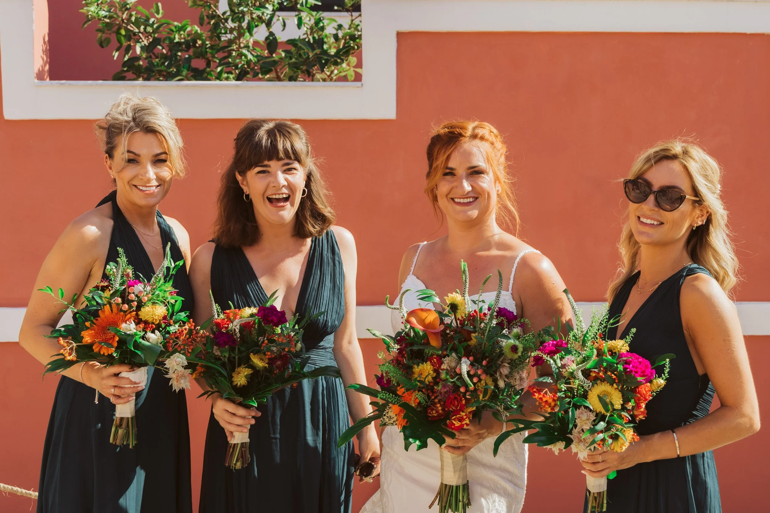 Four women dressed in formal attire standing outdoors holding bouquets of flowers, smiling for a photo.