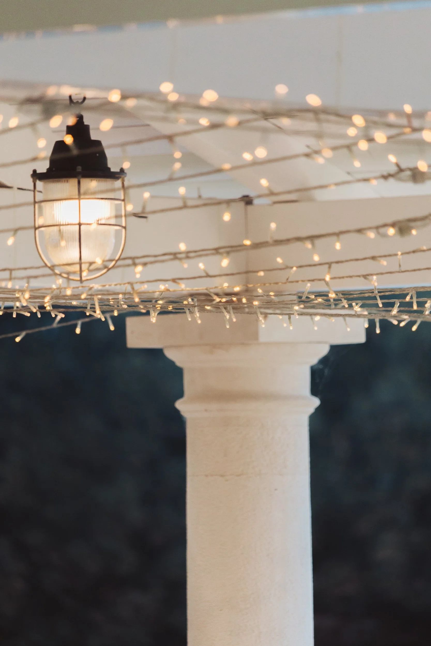 Close-up of outdoor string lights and a lantern hanging from a white porch ceiling, with a white column supporting the structure.