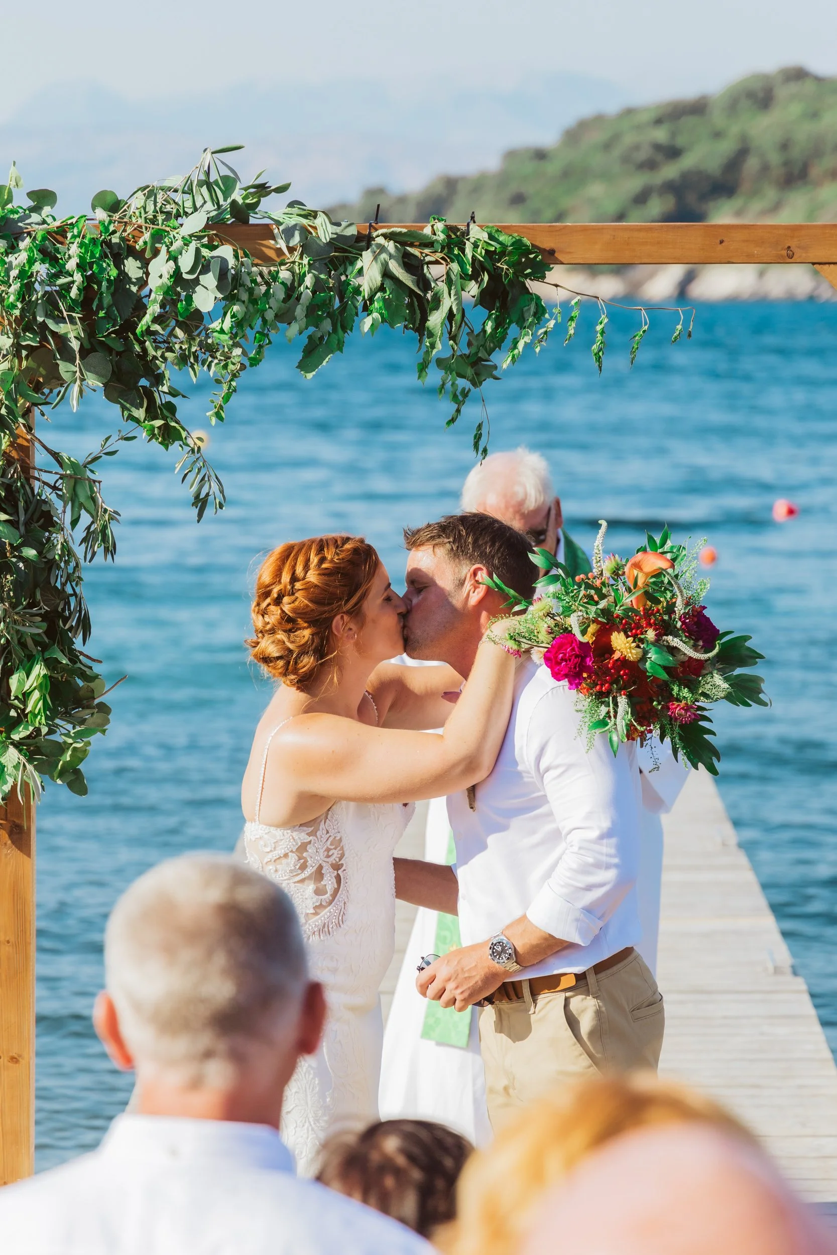 Couple kissing during a wedding ceremony on a dock by the water with guests and a scenic background.