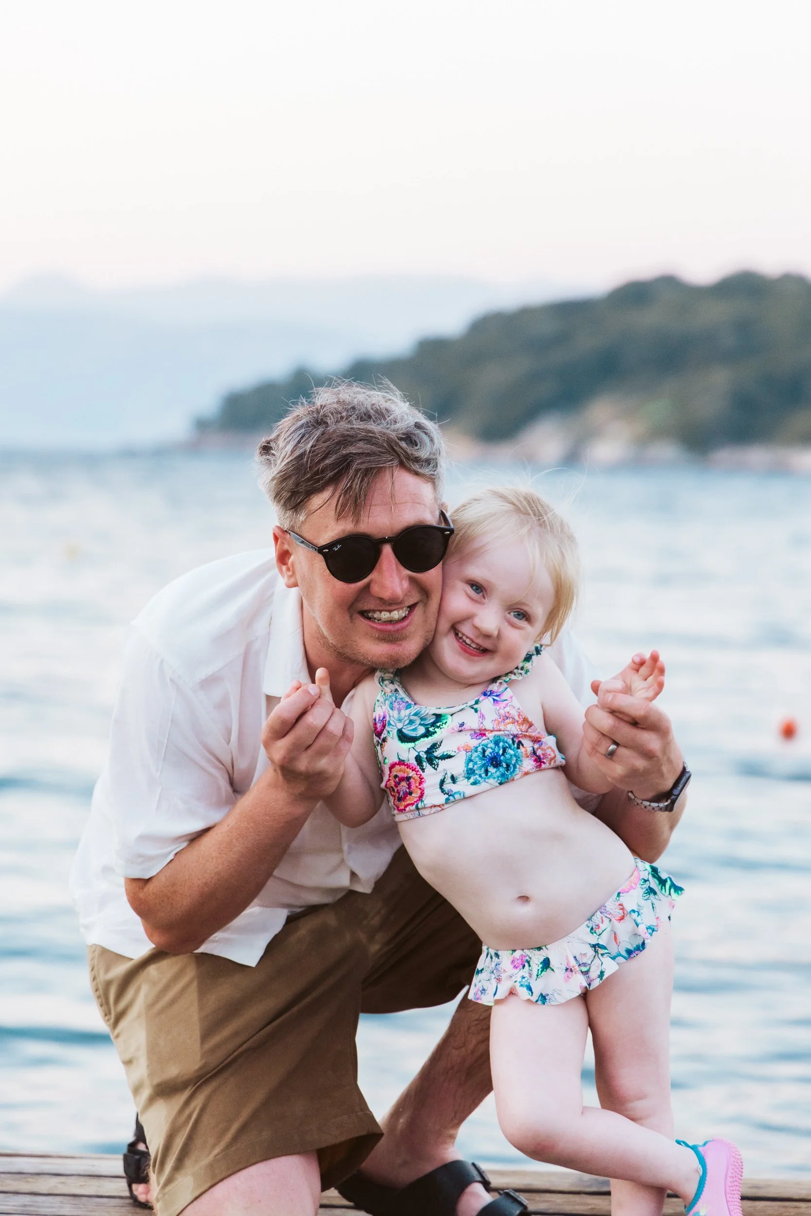A man wearing sunglasses and a white shirt squatting on a dock, holding a smiling young girl in a colorful swimsuit, at a beach with water and distant hills in the background.