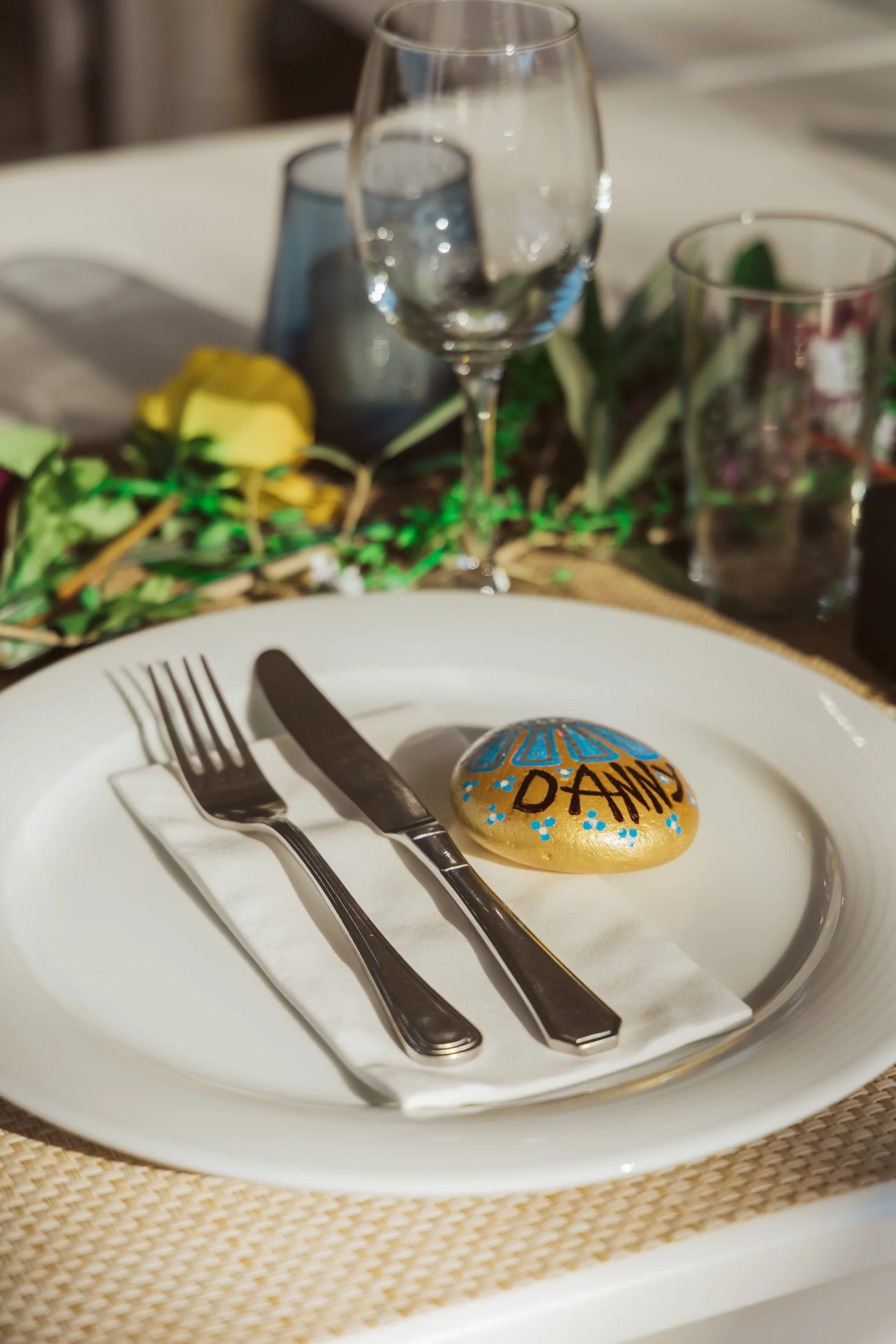 A decorated plate with a painted stone that has the name D'Anne on it, placed on a napkin with a fork and knife, set on a table with wine glasses and greenery in the background.
