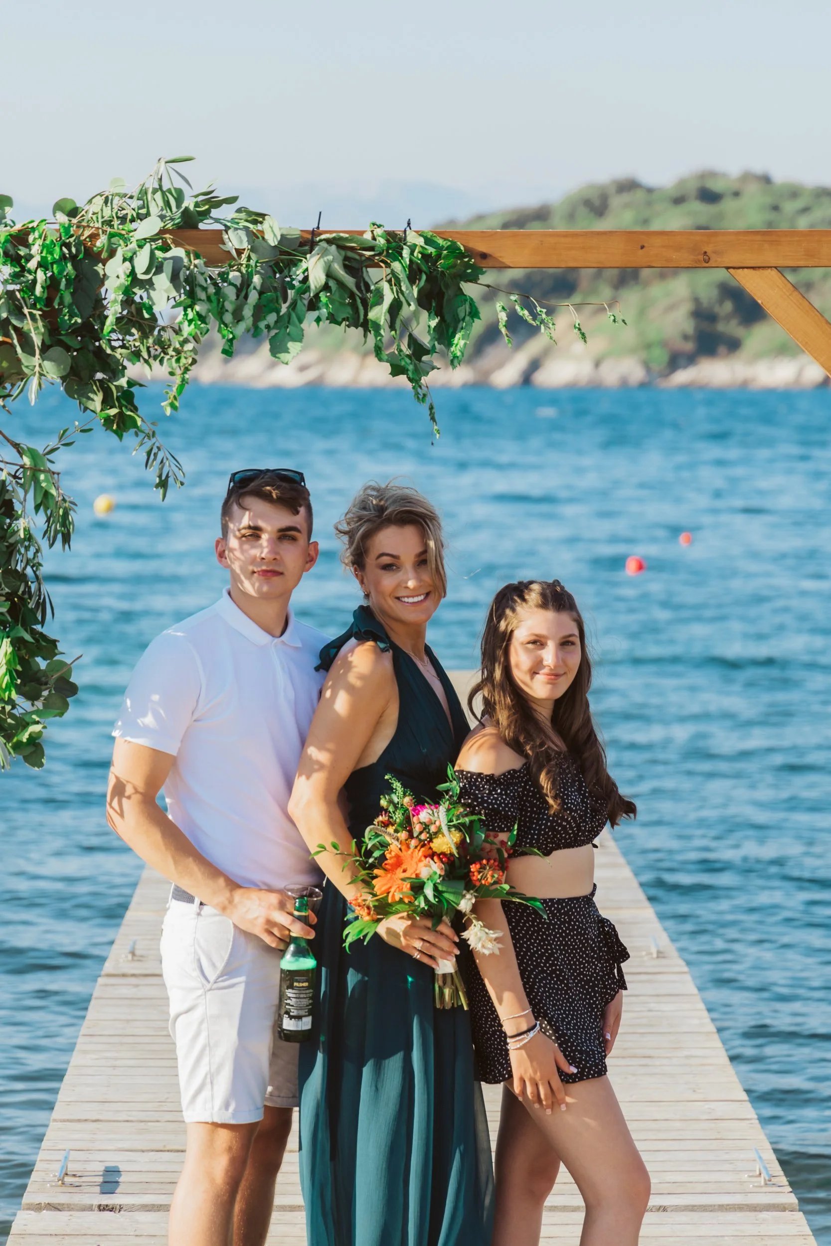 Three people standing on a wooden dock by the water, with hills in the background, under a decorated wooden arch, celebrating a special occasion.