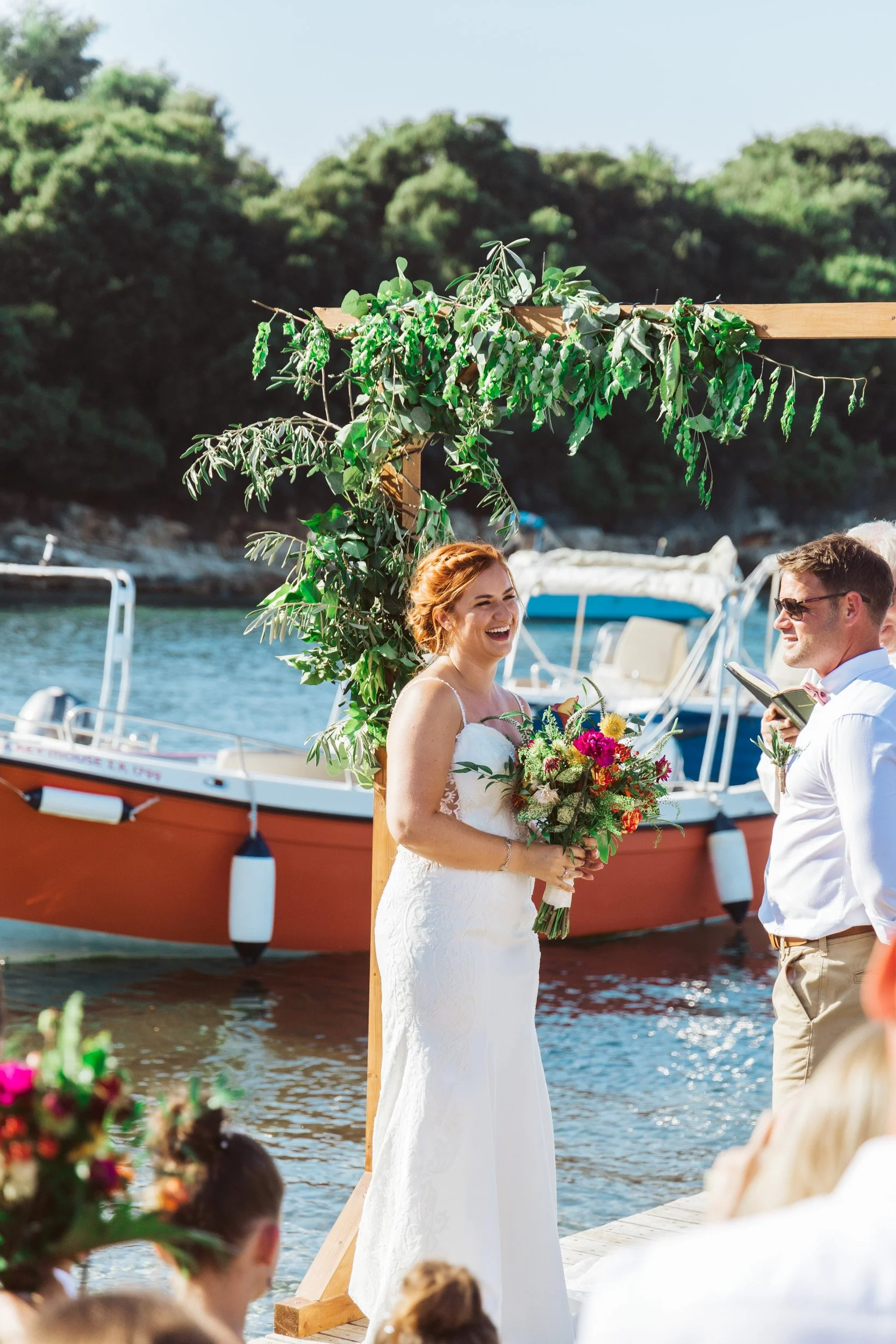 A bride in a white wedding dress holding a bouquet of flowers, standing under a green leafy arch on a dock, with a boat and water in the background.