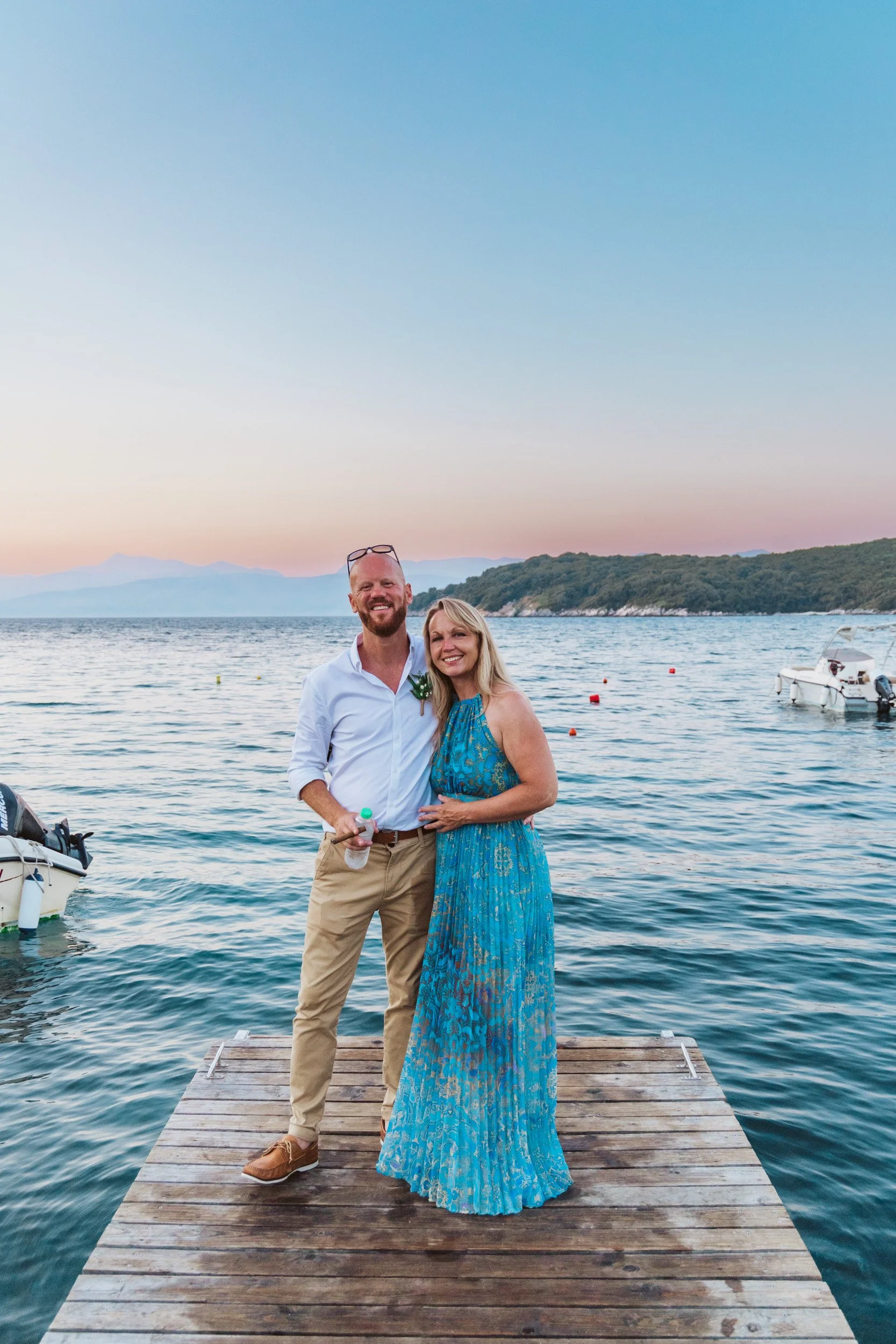 A smiling couple stands on a wooden dock by the water at sunset, with mountains and boats in the background.