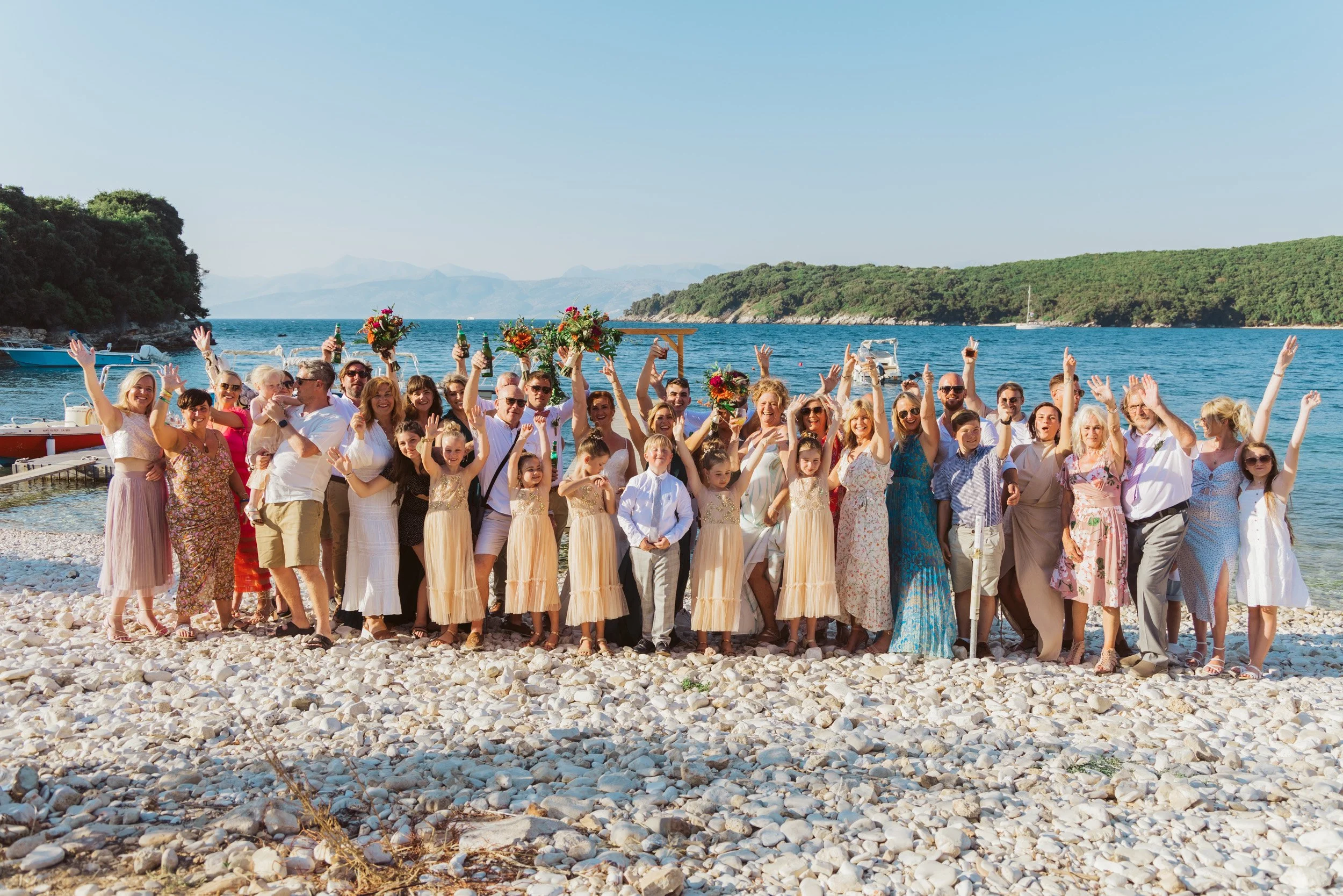 A large group of people celebrating on a pebble beach near a body of water with boats, surrounded by lush green hills and mountains in the background.