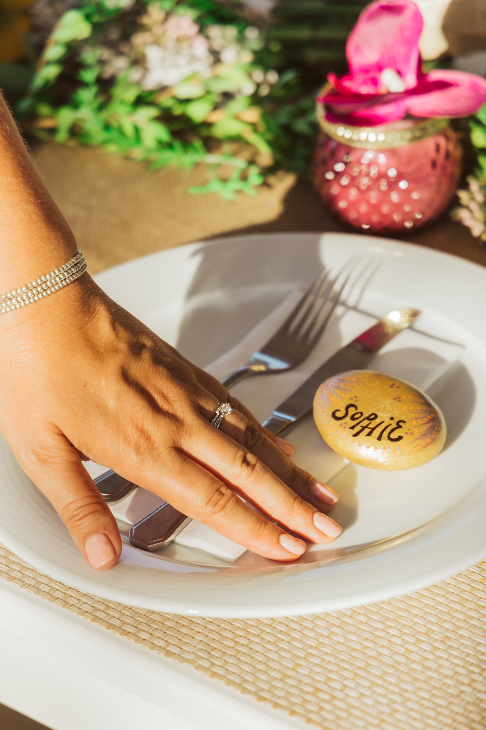 A white dinner plate with a fork and knife, a hand with rings and a bracelet resting on the plate, and a decorated rock with the name 'Sophie' written in black ink. In the background, a colorful vase with pink flowers and greenery.