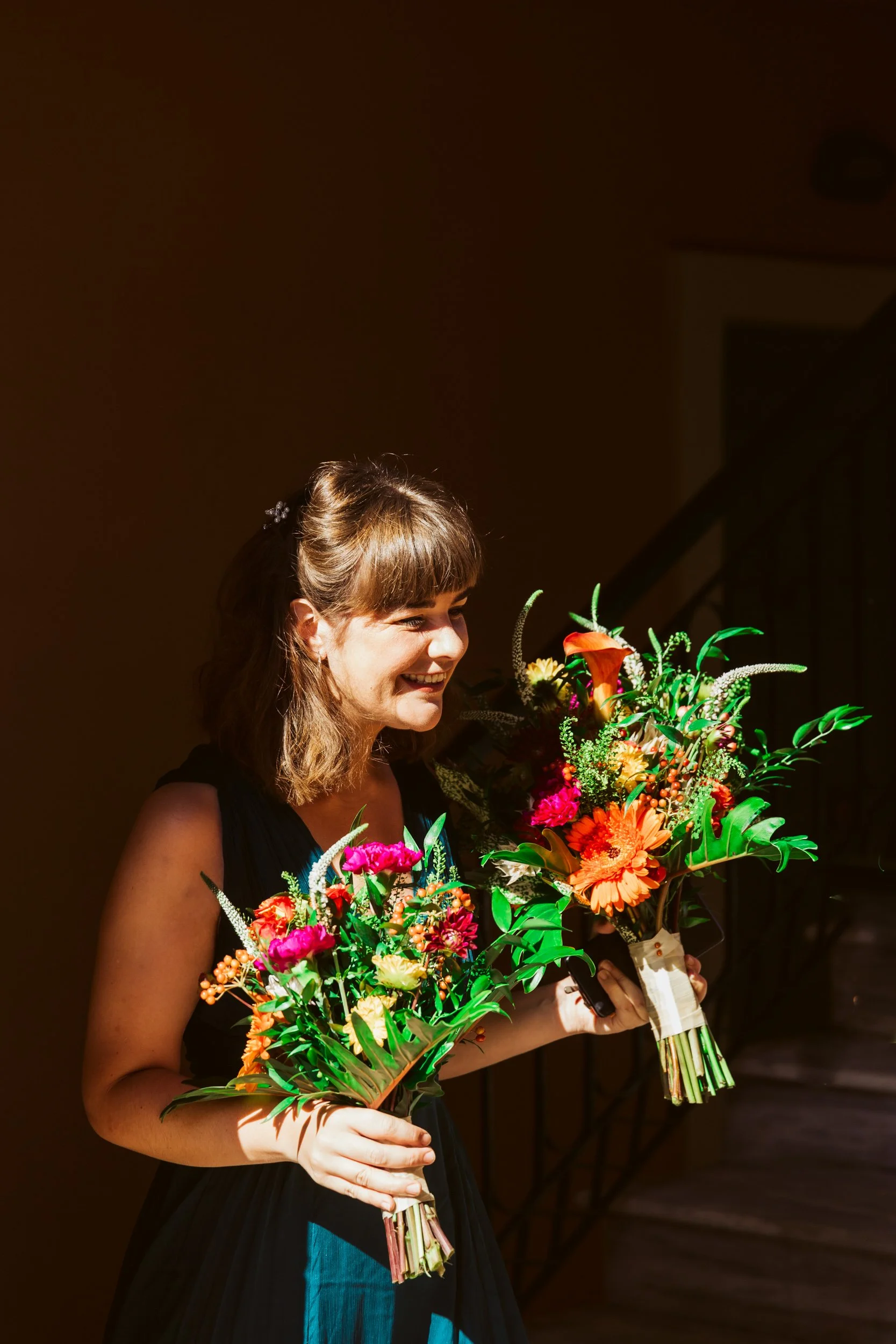 A woman smiling and holding two vibrant flower bouquets, standing indoors with warm lighting.