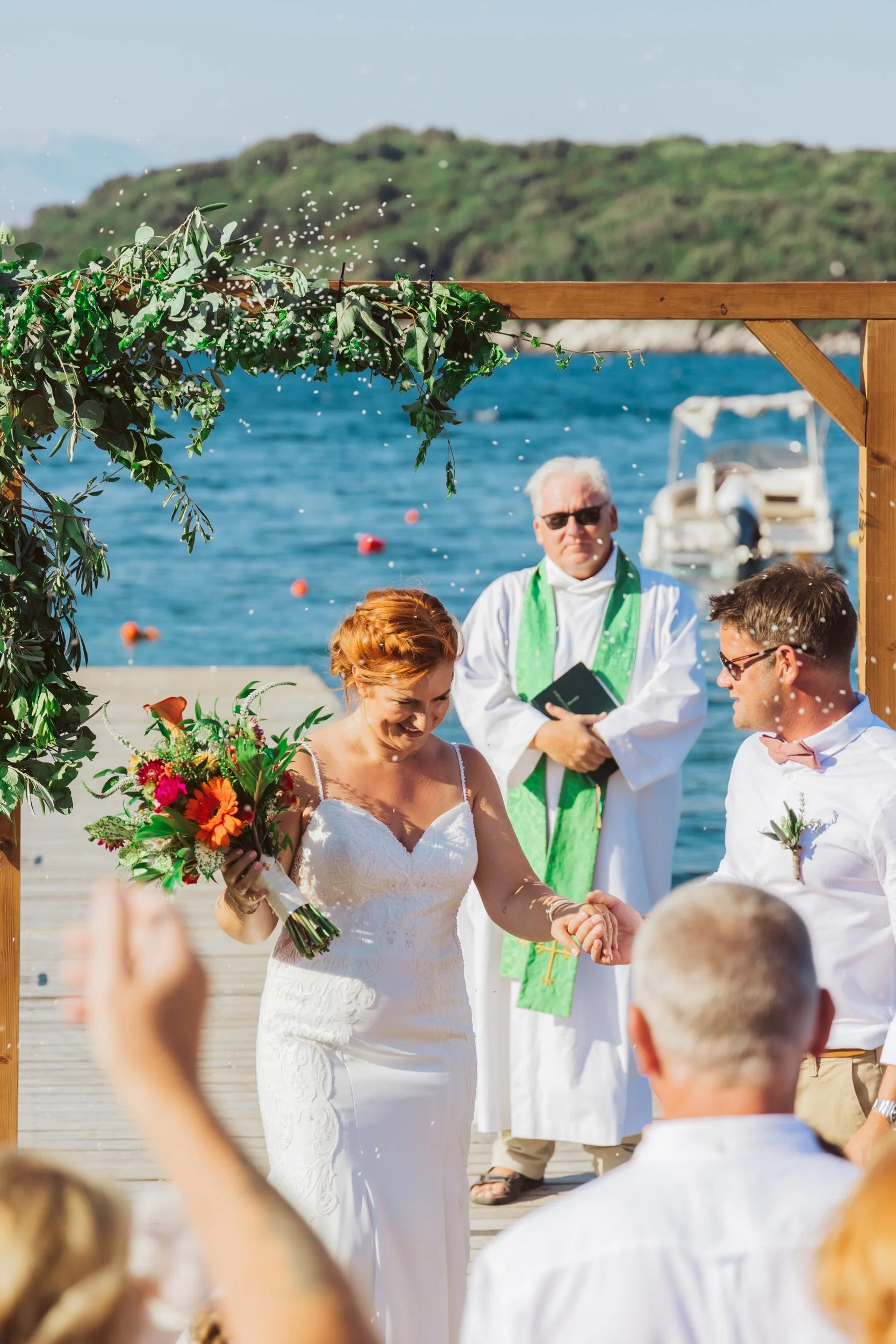 A bride and groom holding hands during their outdoor wedding ceremony on a dock by the water, with an officiant standing behind them and guests capturing the moment.