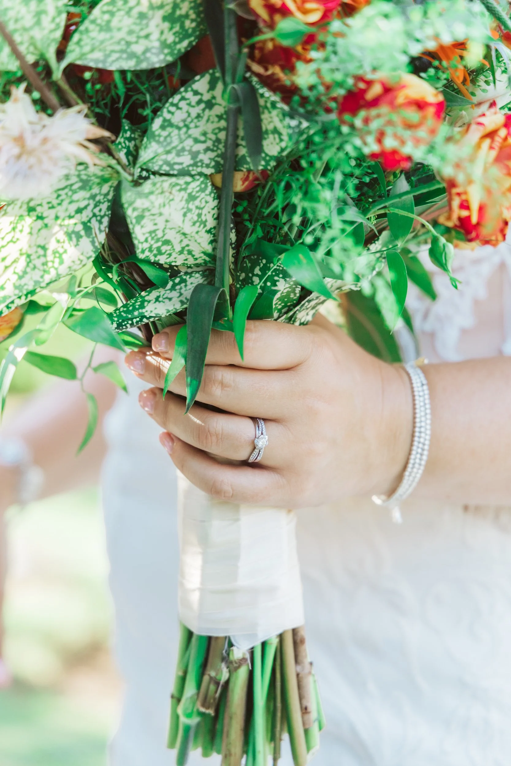 Close-up of a woman's hand holding a bouquet of assorted flowers and greenery, wearing a diamond engagement ring and a pearl bracelet.