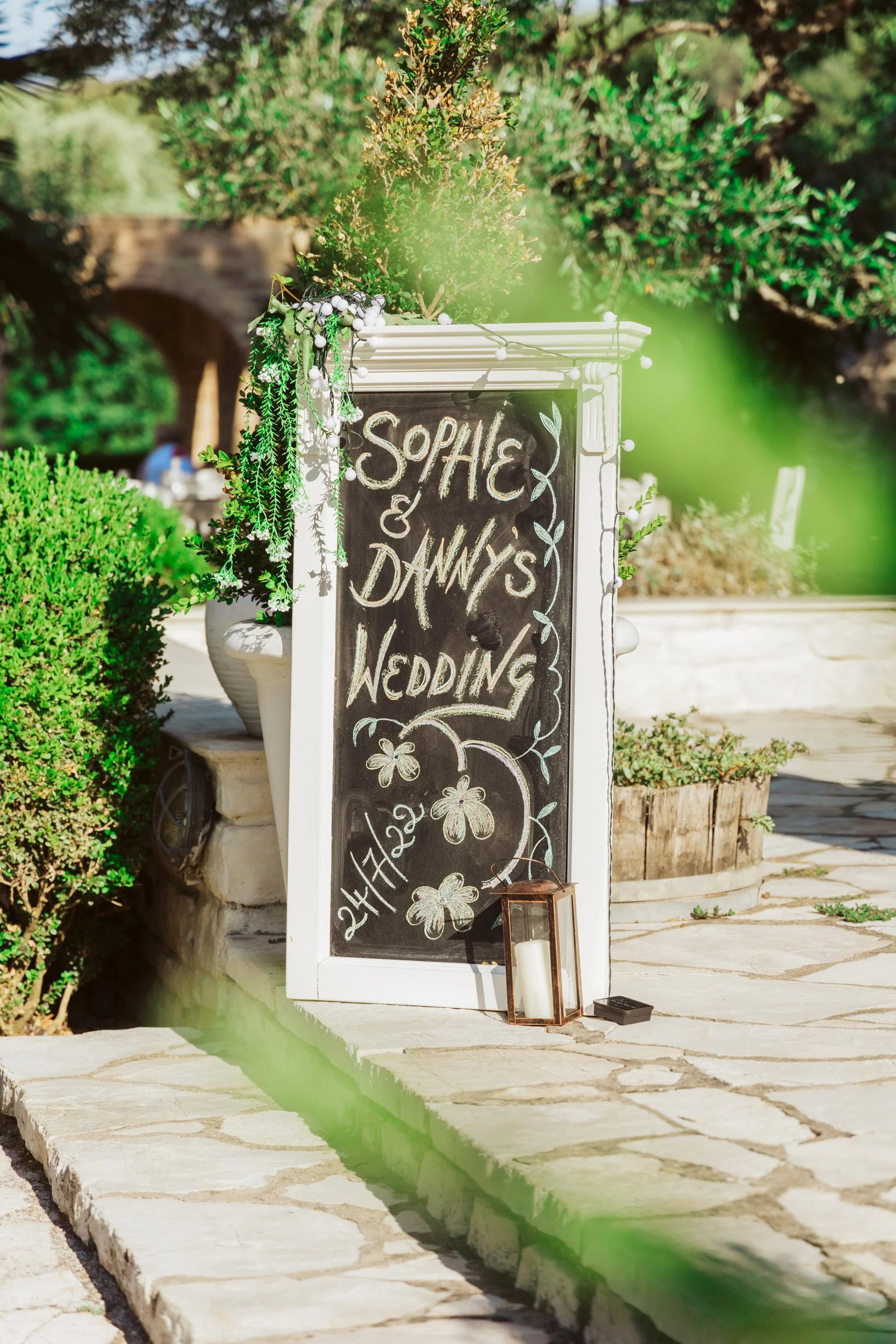 A blackboard sign with white and colorful chalk writing announcing Sophie and Danny's wedding, scheduled for April 22, 2023. The sign is decorated with flowers and greenery, and there is a candle lantern placed at the base. The setting appears to be 