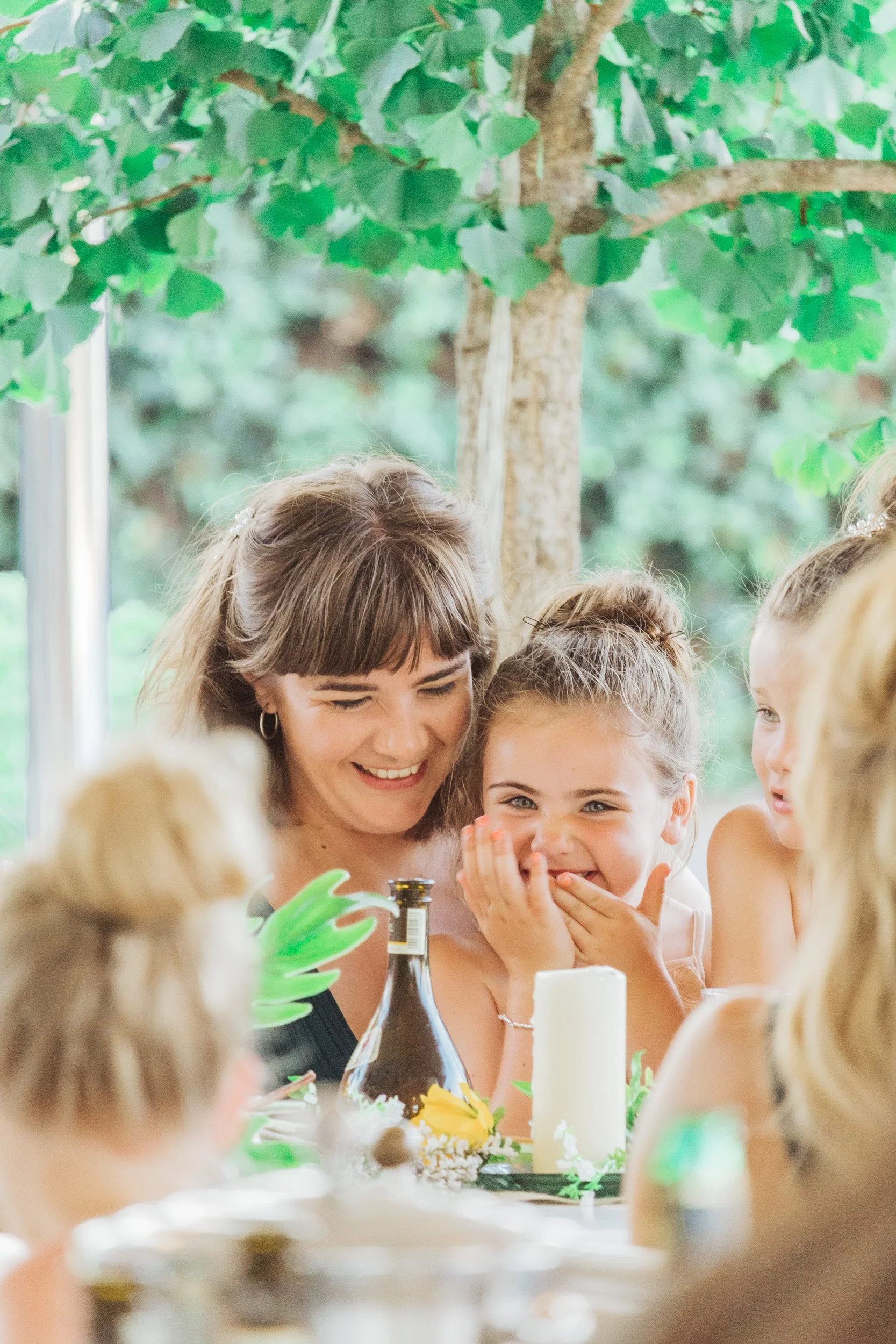 A woman and a girl sharing a laugh at an outdoor party with children, candles, and greenery.