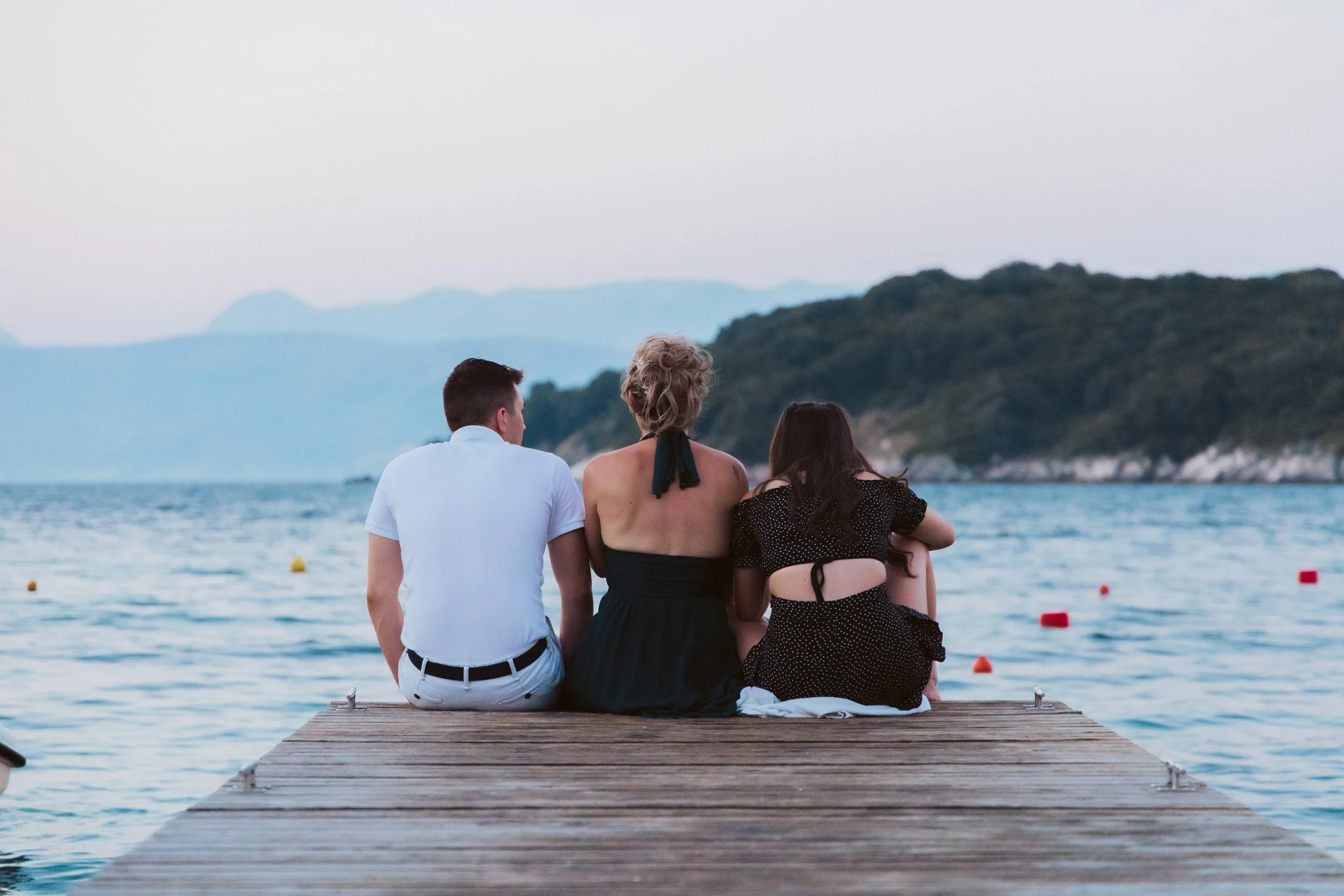 Three people sit on a wooden dock overlooking a body of water with distant mountains and trees in the background, during sunset or dusk.
