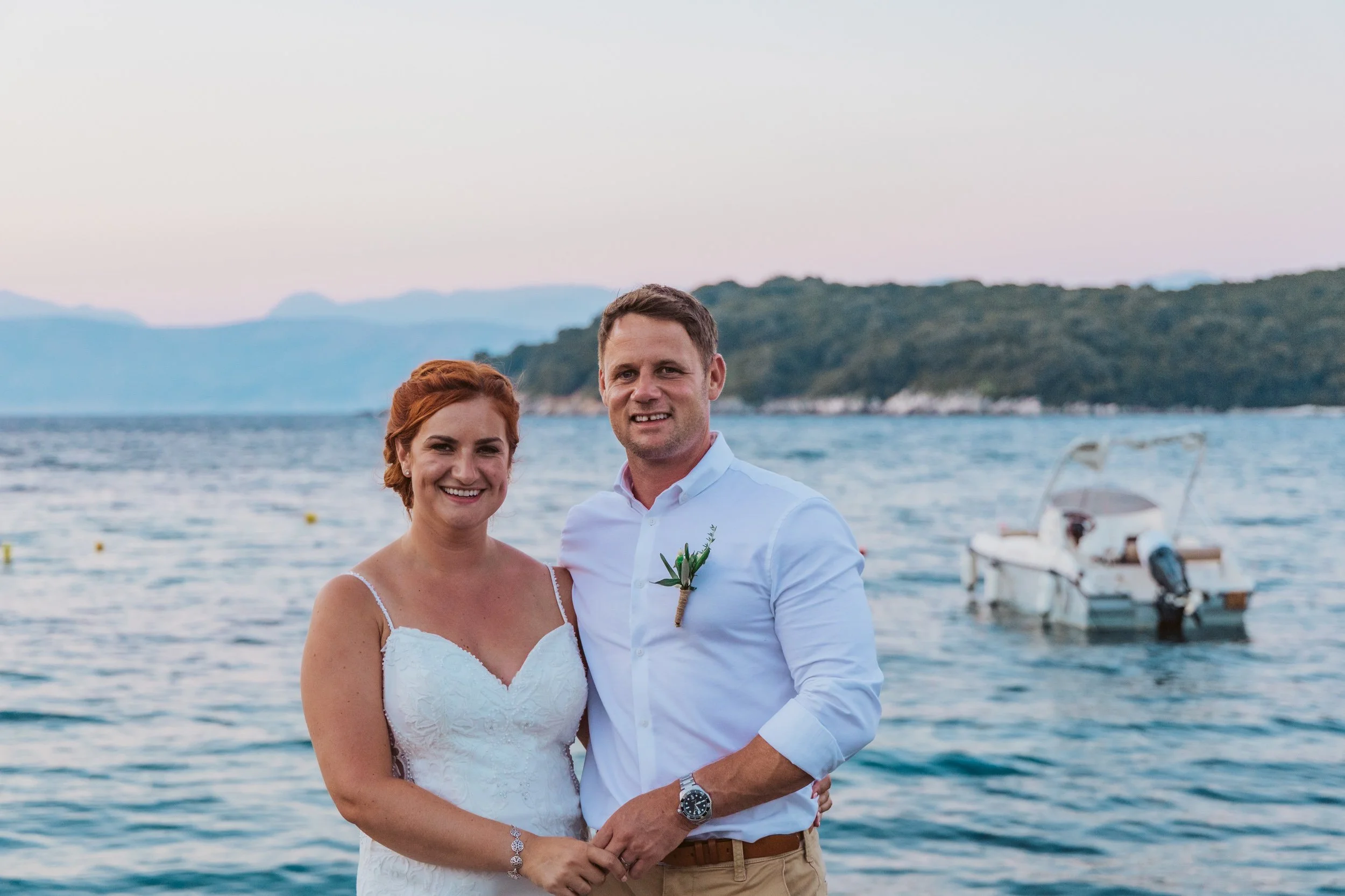 A smiling bride and groom standing by a body of water with mountains in the background, wedding attire, and a boat nearby.