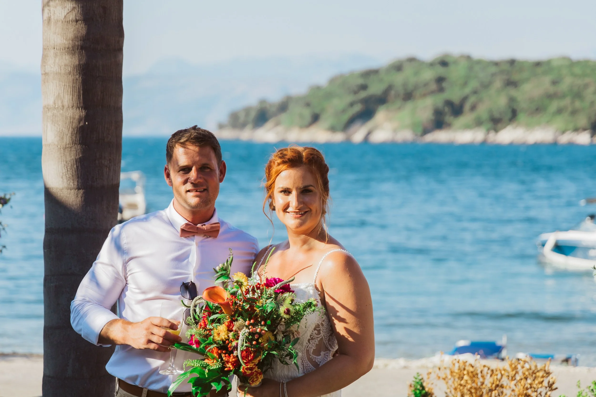 A bride and groom standing together on a beach with water, boats, and a hillside in the background. The man is holding a glass and wearing a white shirt with a pink bow tie, the woman is holding a colorful bouquet and wearing a white dress.