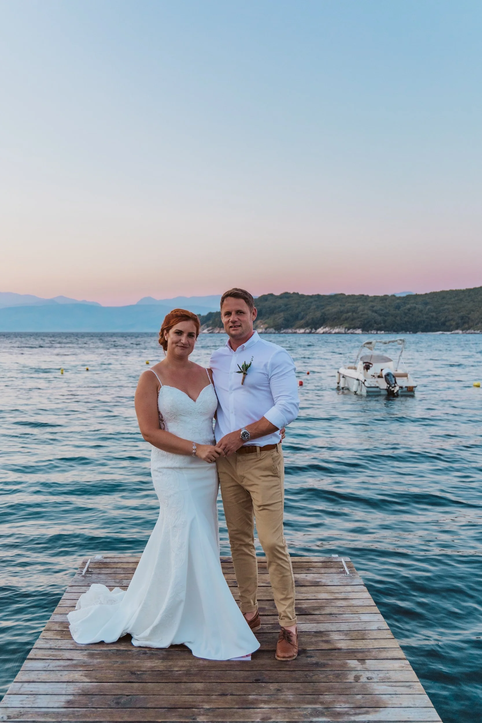 A bride and groom standing on a wooden dock by the water at sunset, with a boat and distant hills in the background.