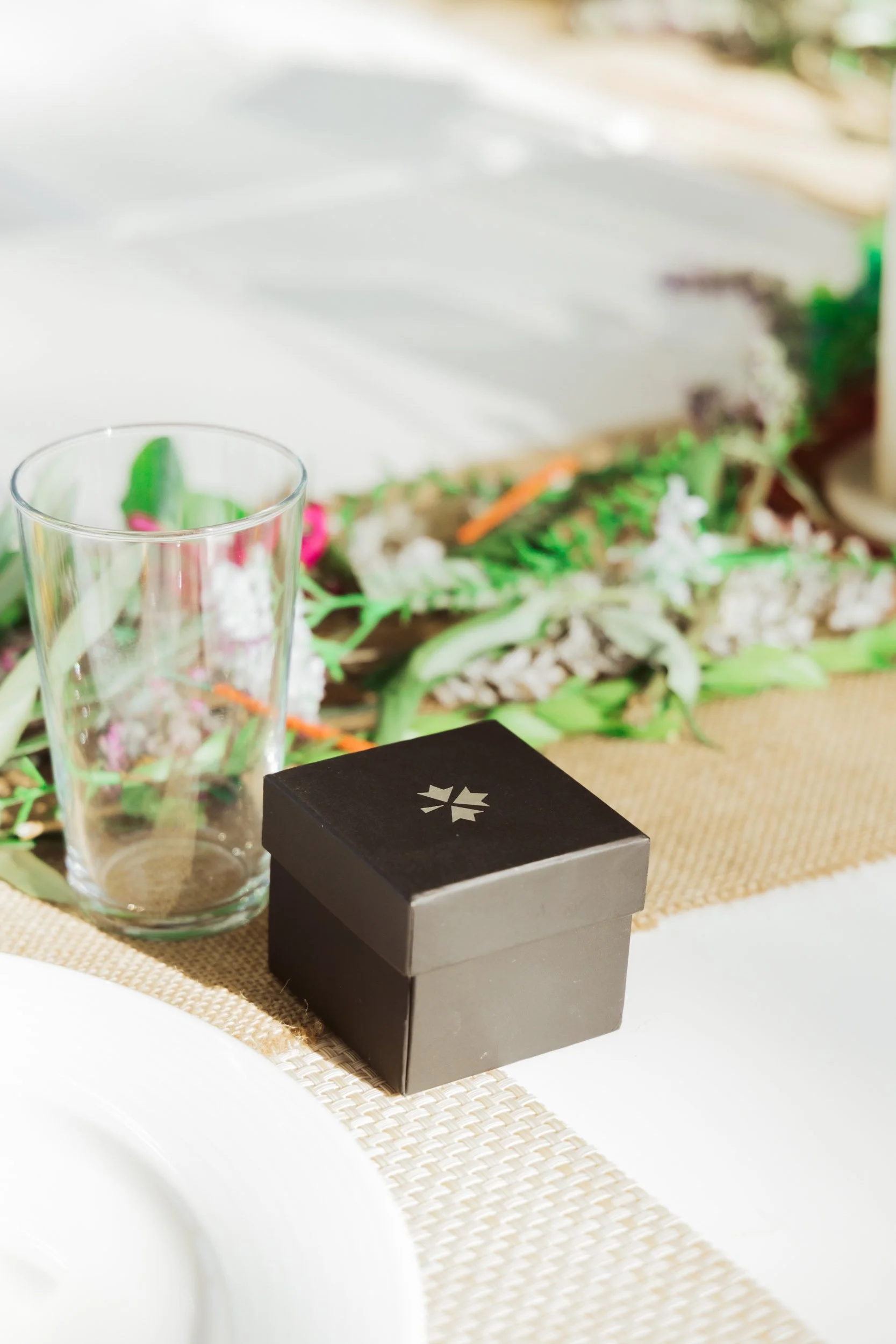 A black gift box with a white logo, an empty glass, and a floral centerpiece on a table.