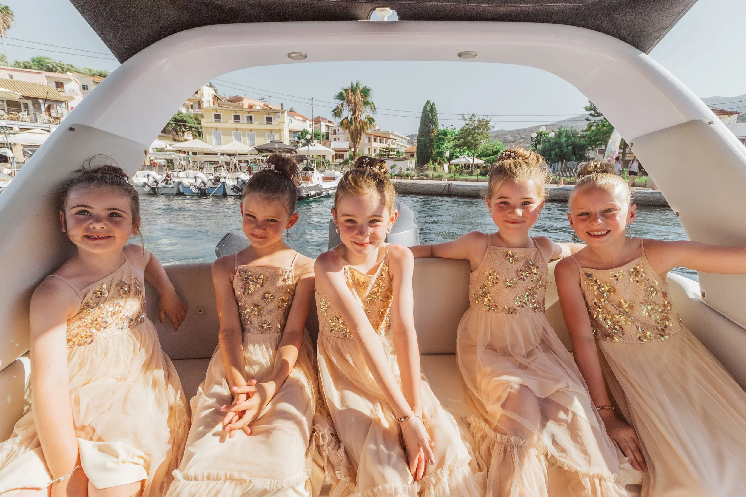 Six young girls in matching peach-colored dresses with gold embellishments sitting on a boat with water and a marina in the background.