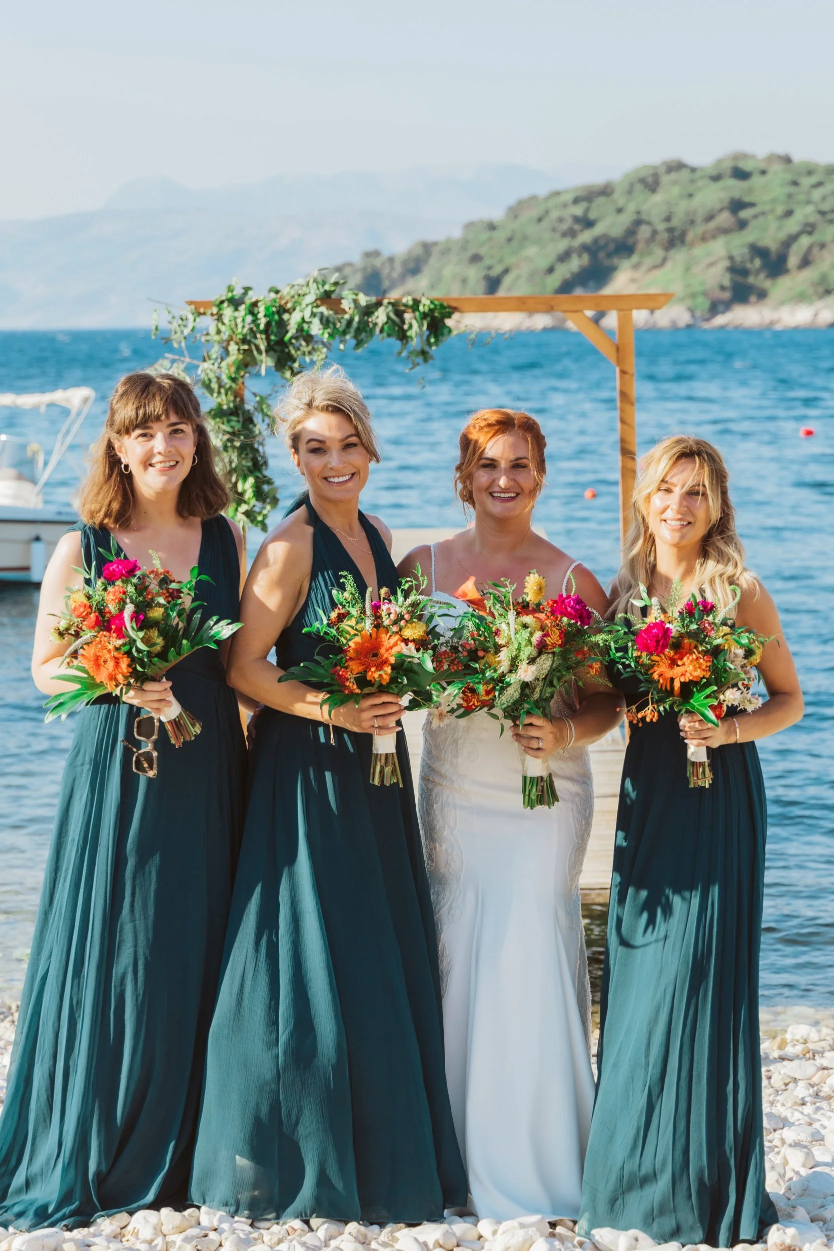 Four women in formal dresses standing on a beach with water and mountains in the background. The woman second from right is wearing a white dress, indicating she may be the bride. The other three women are holding colorful bouquets and are likely bri