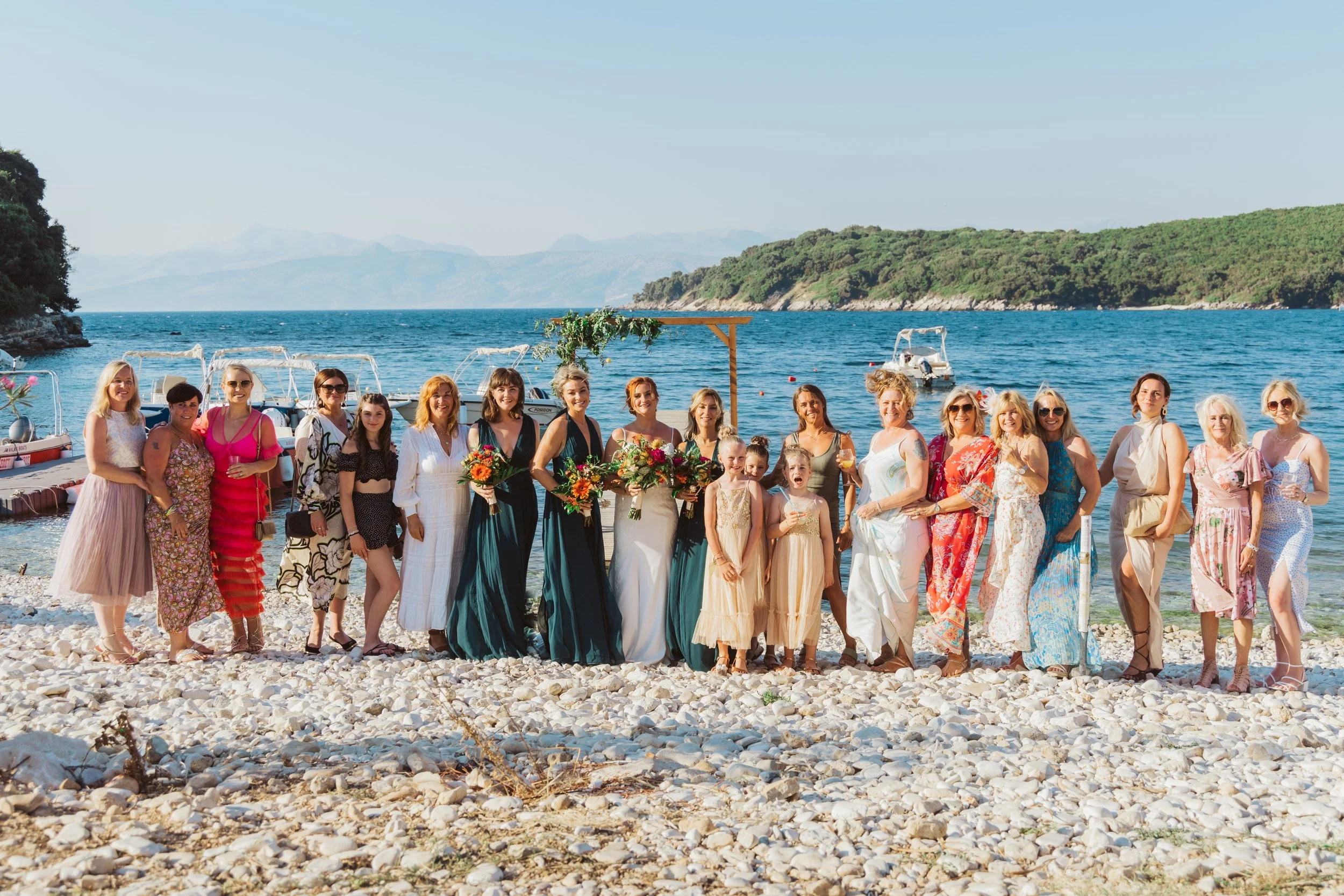 A group of women and children on a rocky beach near the water, celebrating a wedding with a wooden arch, some holding bouquets, and dressed in bright, summery attire.