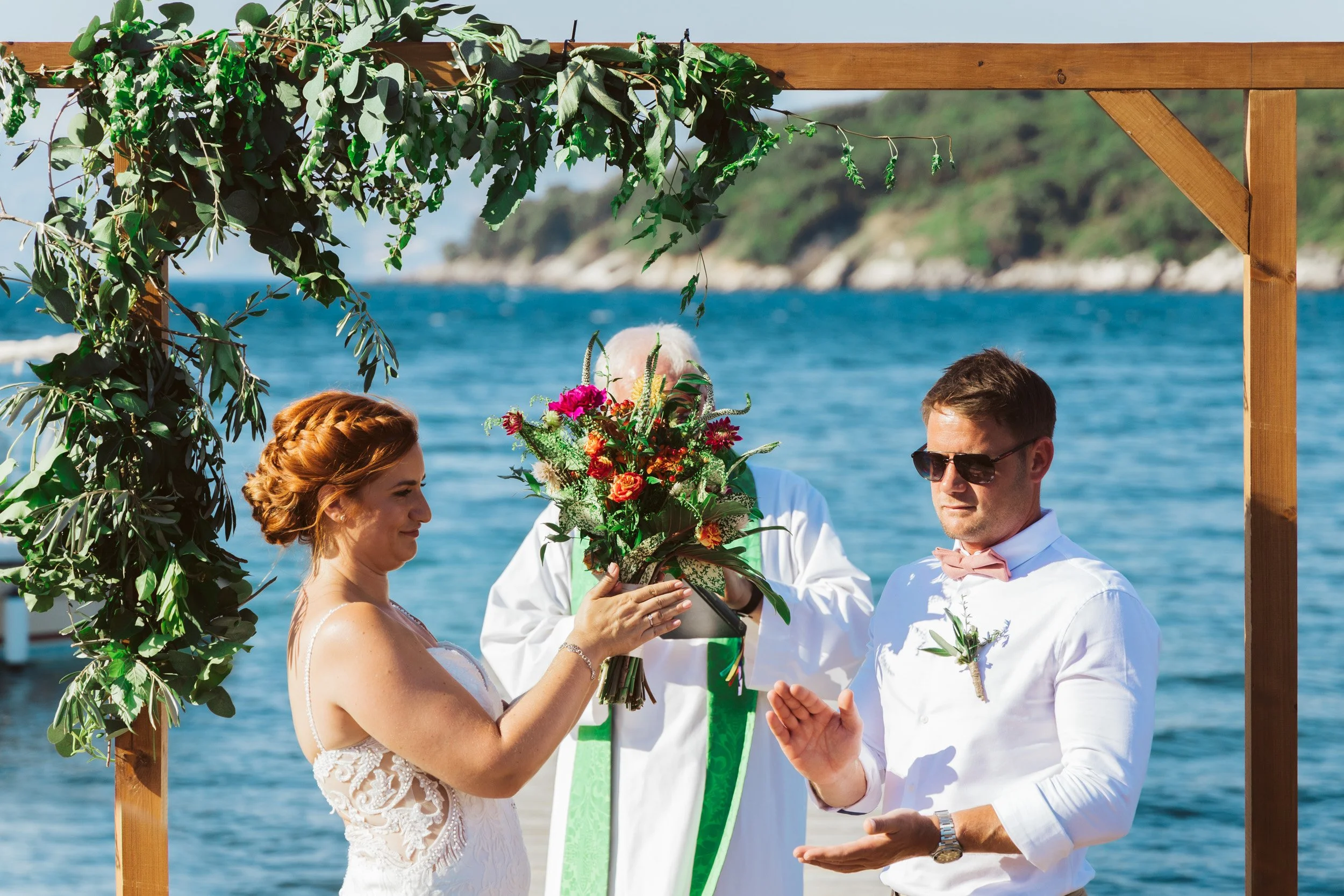 A couple at their wedding ceremony by the water, with a celebration taking place under a wooden arch decorated with green leaves. The bride is holding a bouquet and the groom is wearing sunglasses and a boutonnière.