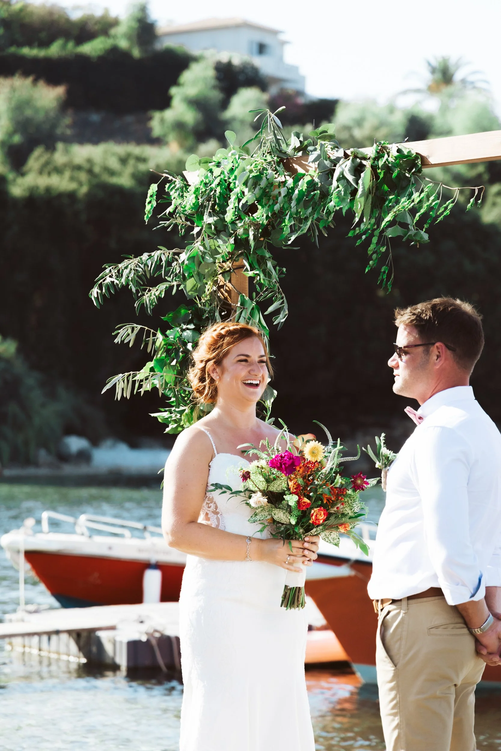 A bride and groom stand outdoors near the water, facing each other during their wedding ceremony. The bride holds a colorful bouquet and is smiling, while the groom looks at her. There is greenery and a boat in the background.