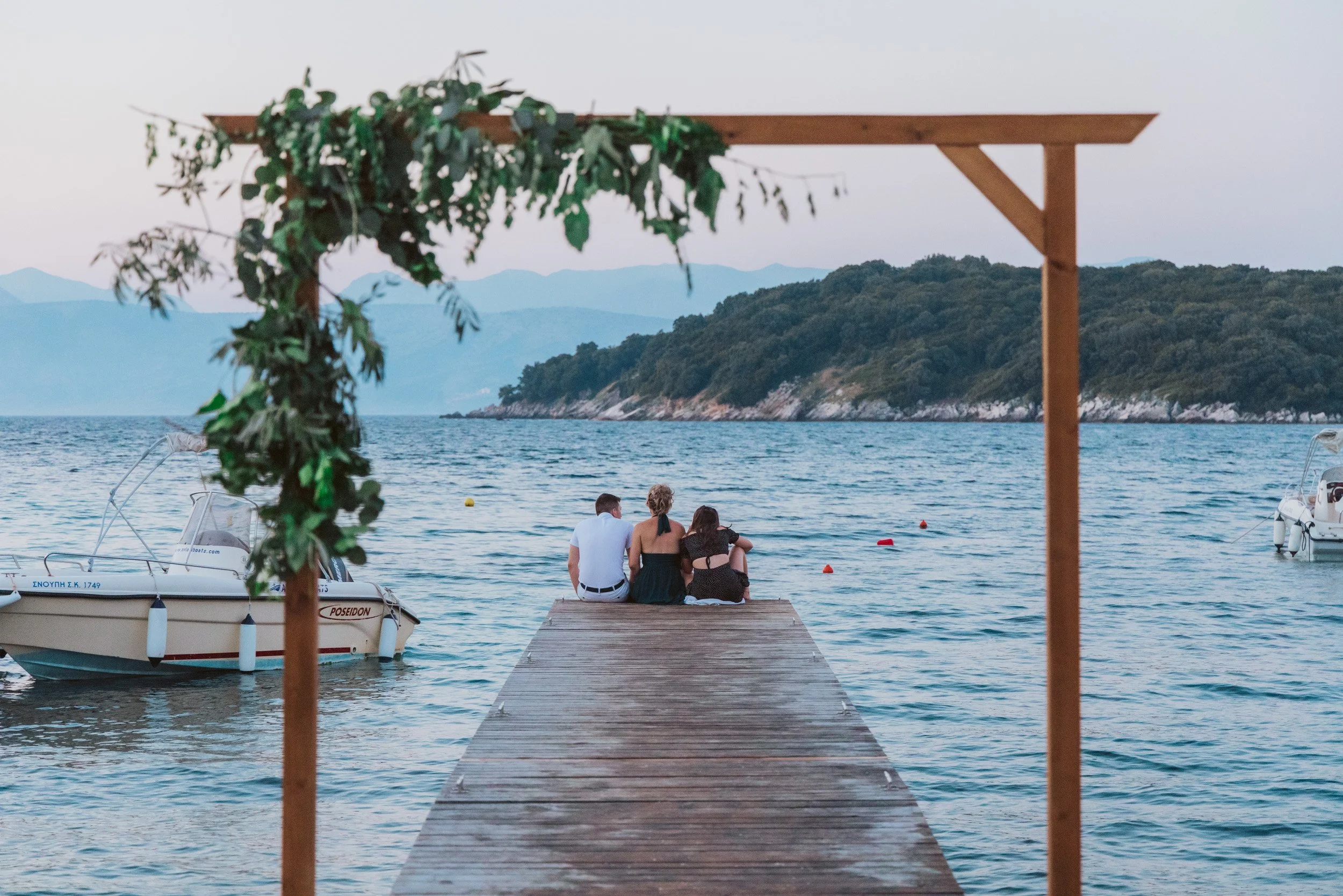 Three people sitting on a pier at a marina, with boats on the water and mountains in the background, framed by a wooden arch with green foliage.