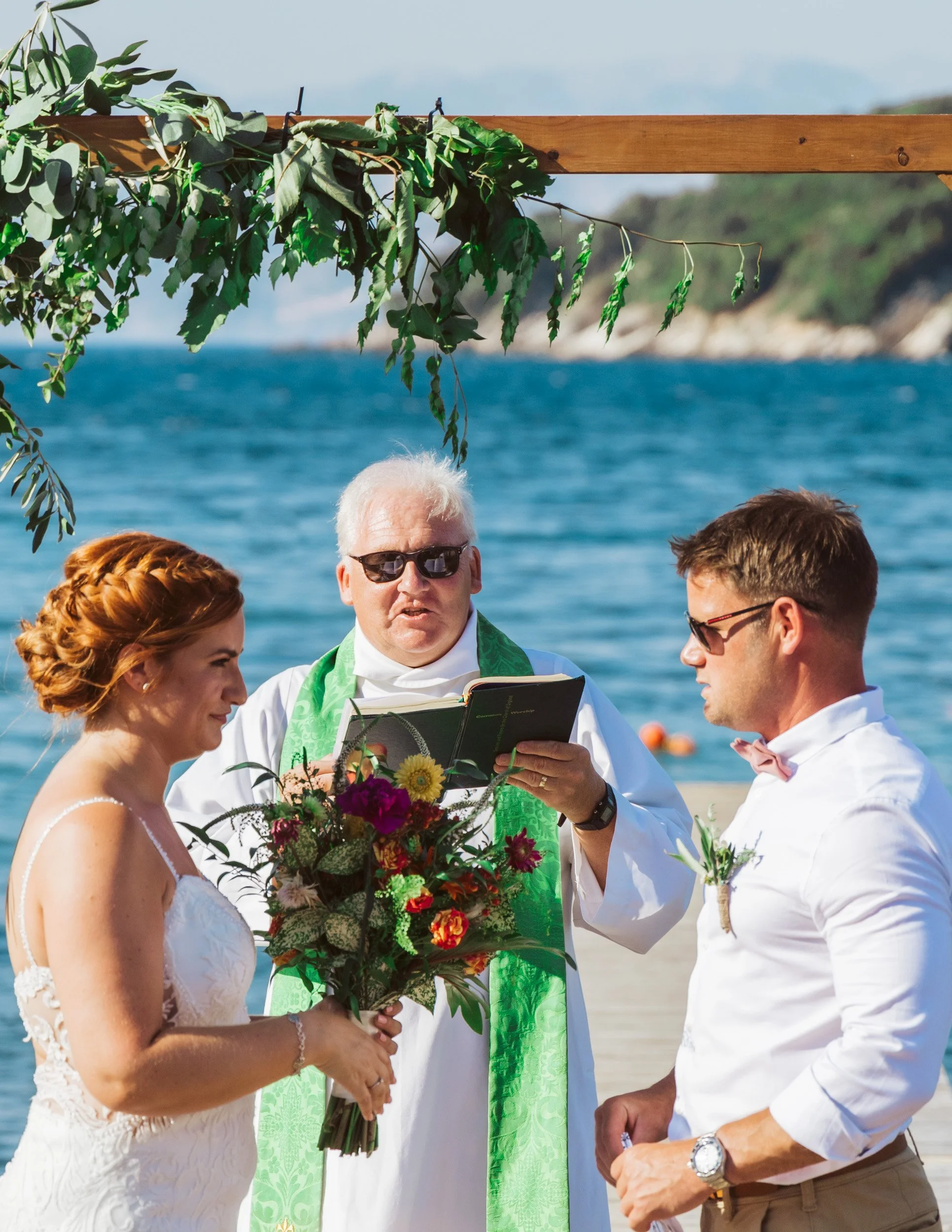 A bride and groom getting married outdoors on a beach with an officiant. The bride has red hair styled in curls, wearing a white lace wedding dress, and holding a bouquet. The groom has short brown hair, wearing a white shirt with rolled-up sleeves a
