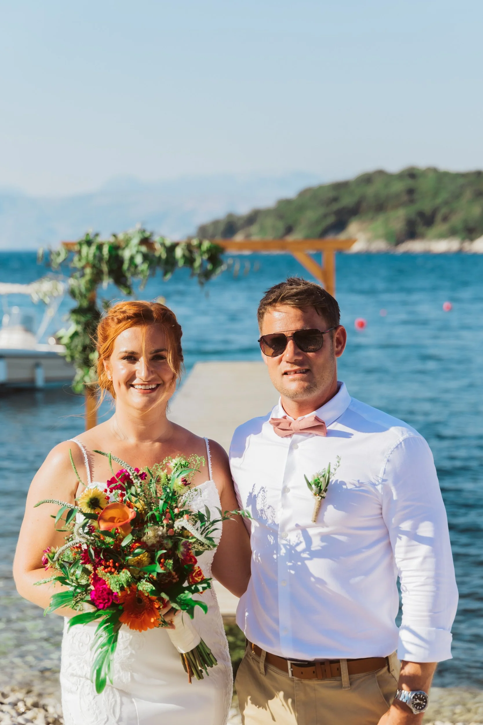A happy bride and groom stand outdoors near a body of water, with the bride holding a colorful bouquet of flowers, under a wooden arch decorated with greenery, during their wedding celebration on a sunny day.