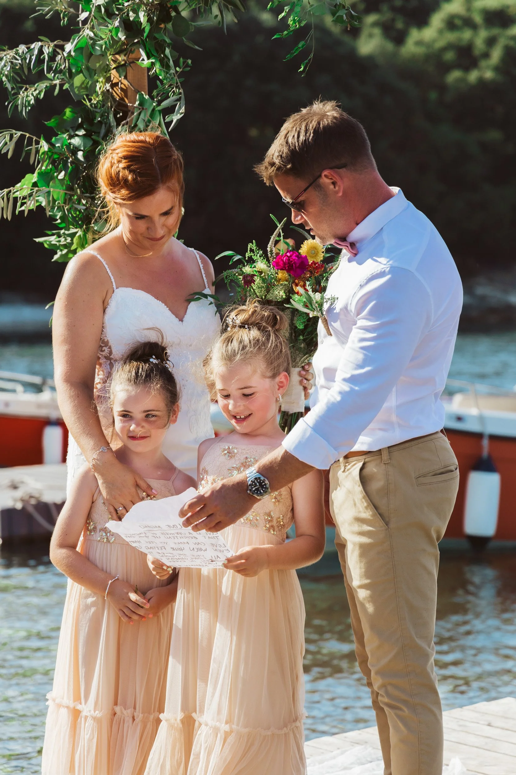 A wedding ceremony outdoors by the water with a bride, groom, and two young girls. The groom is presenting a paper to the girls, while the bride holds a colorful bouquet. The scene is sunny and surrounded by greenery.