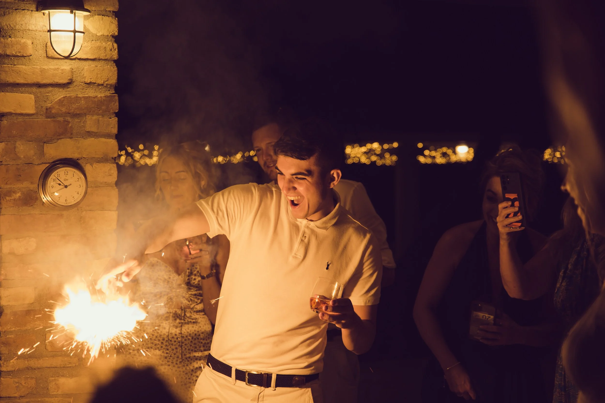 People celebrating at a nighttime gathering, with a young man holding a glass and playing with sparklers next to an outdoor brick wall with a clock showing 11:24, and city lights in the distance.