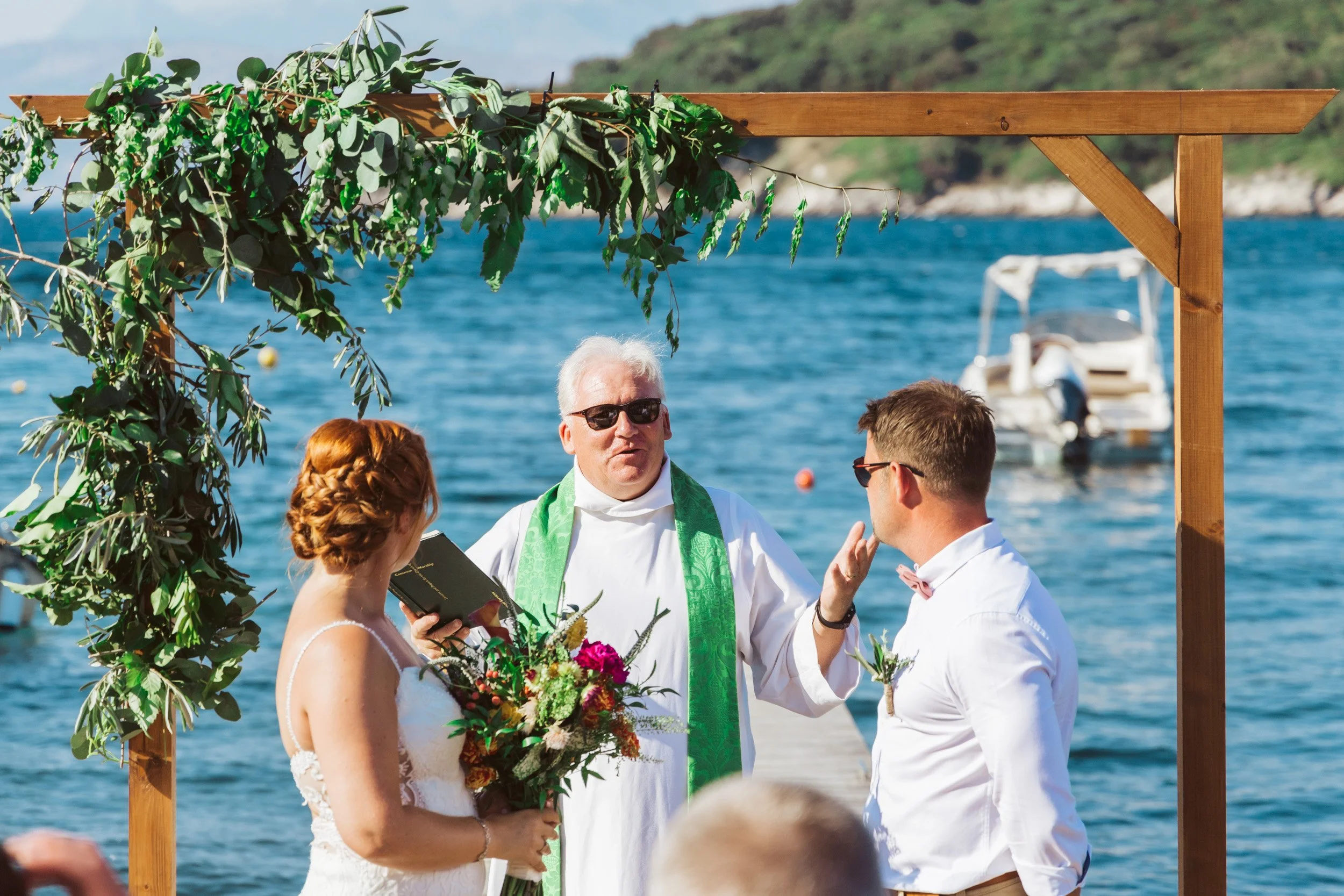 A wedding ceremony taking place on a dock by the water with a priest conducting the vows, a bride holding a bouquet, and a groom in sunglasses, with a boat and blue water in the background.