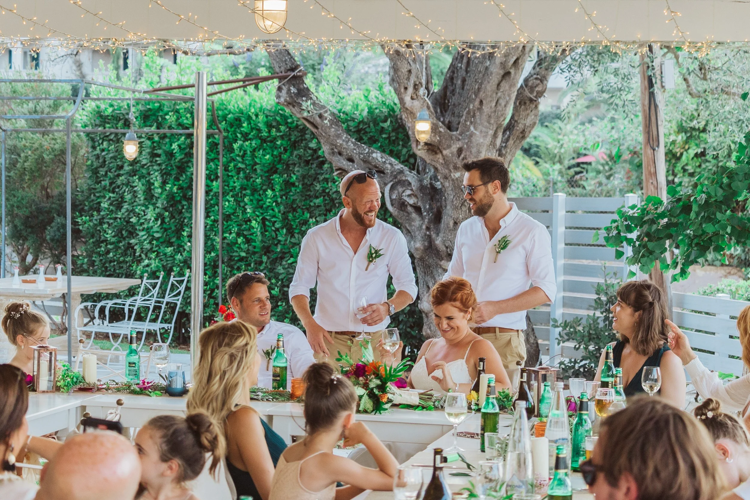 A group of people gathered around tables outdoors for a celebration. Two men are standing and talking, while others, including women and children, are seated and smiling. The setting is decorated with string lights, flowers, and greenery.
