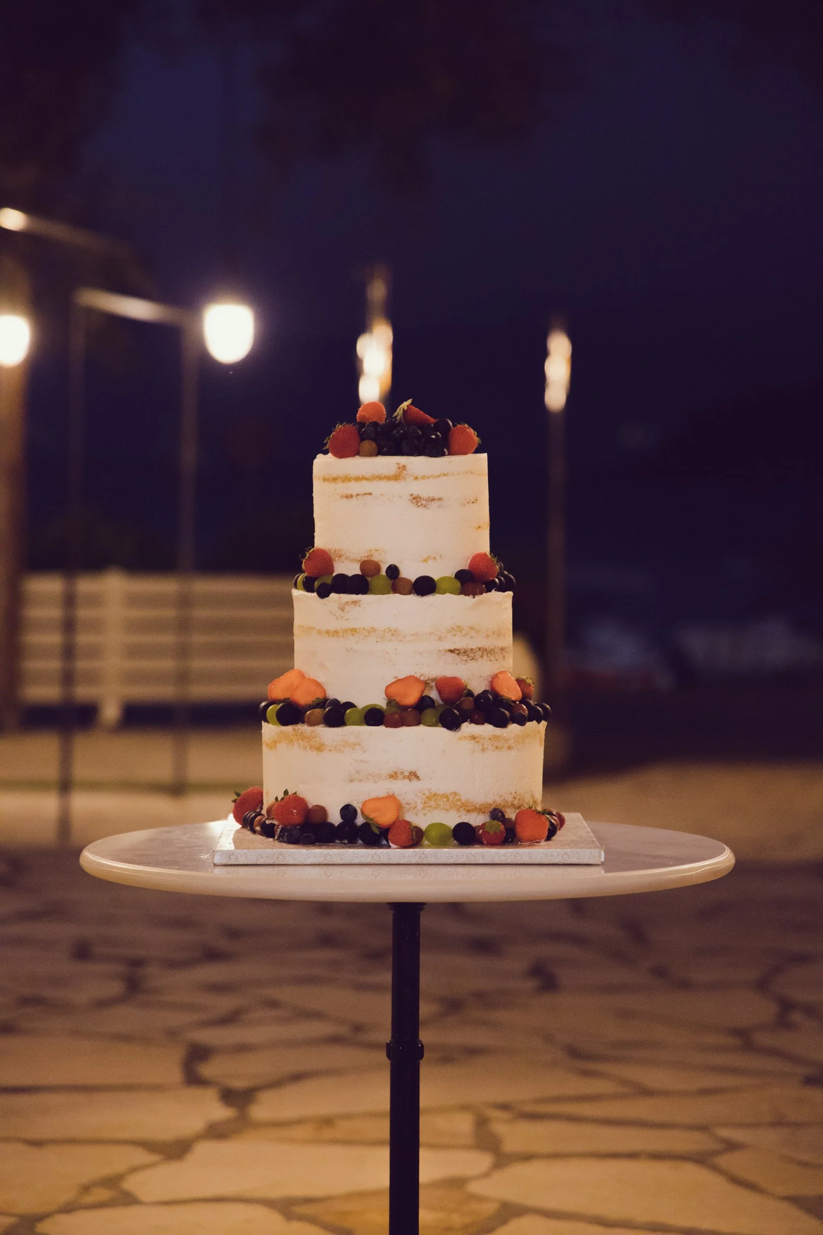 Three-tiered white cake decorated with fresh berries and small flowers, placed on a round table outdoors at night.