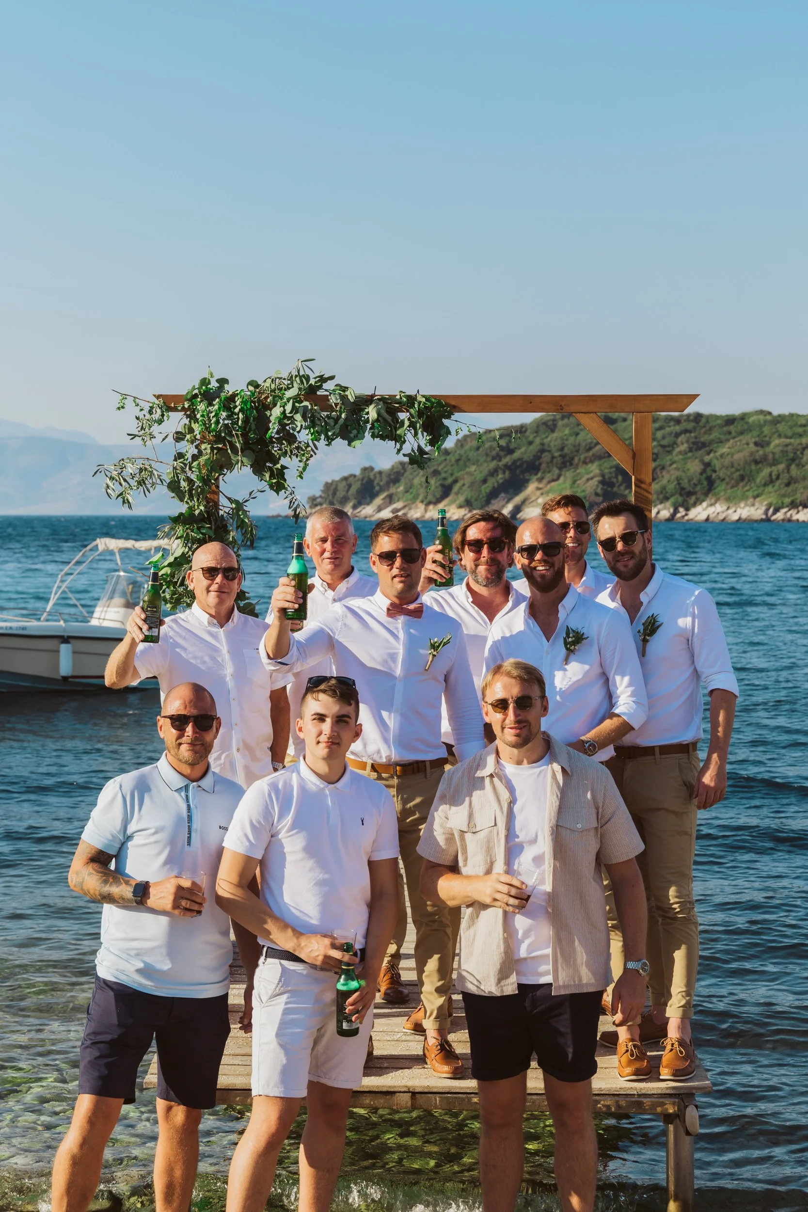 Group of men celebrating on a dock by the water, some holding drinks, with a boat and scenic landscape in the background.
