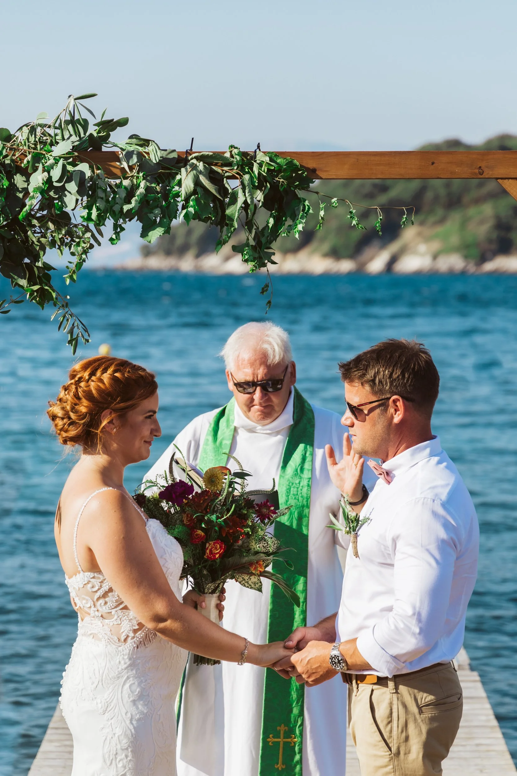 A couple getting married outdoors on a dock by the water, with an officiant present. The bride has red hair and wears a white wedding dress, holding a bouquet of flowers. The groom has dark hair and wears a white shirt with a bow tie and khaki pants.