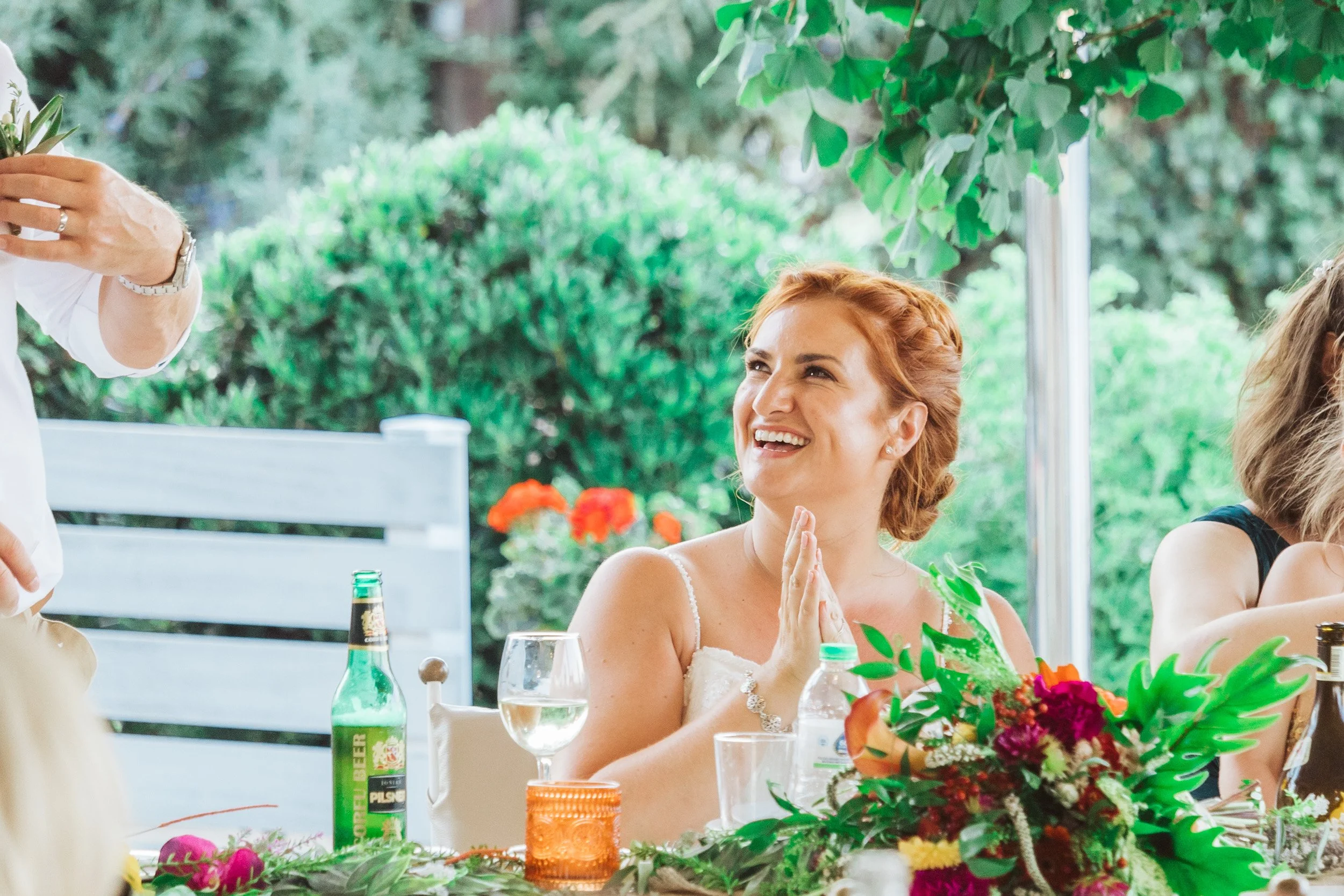 Woman with red hair smiling and holding her hands together at a table during an outdoor celebration, possibly a wedding, with flowers, drinks, and greenery in the background.