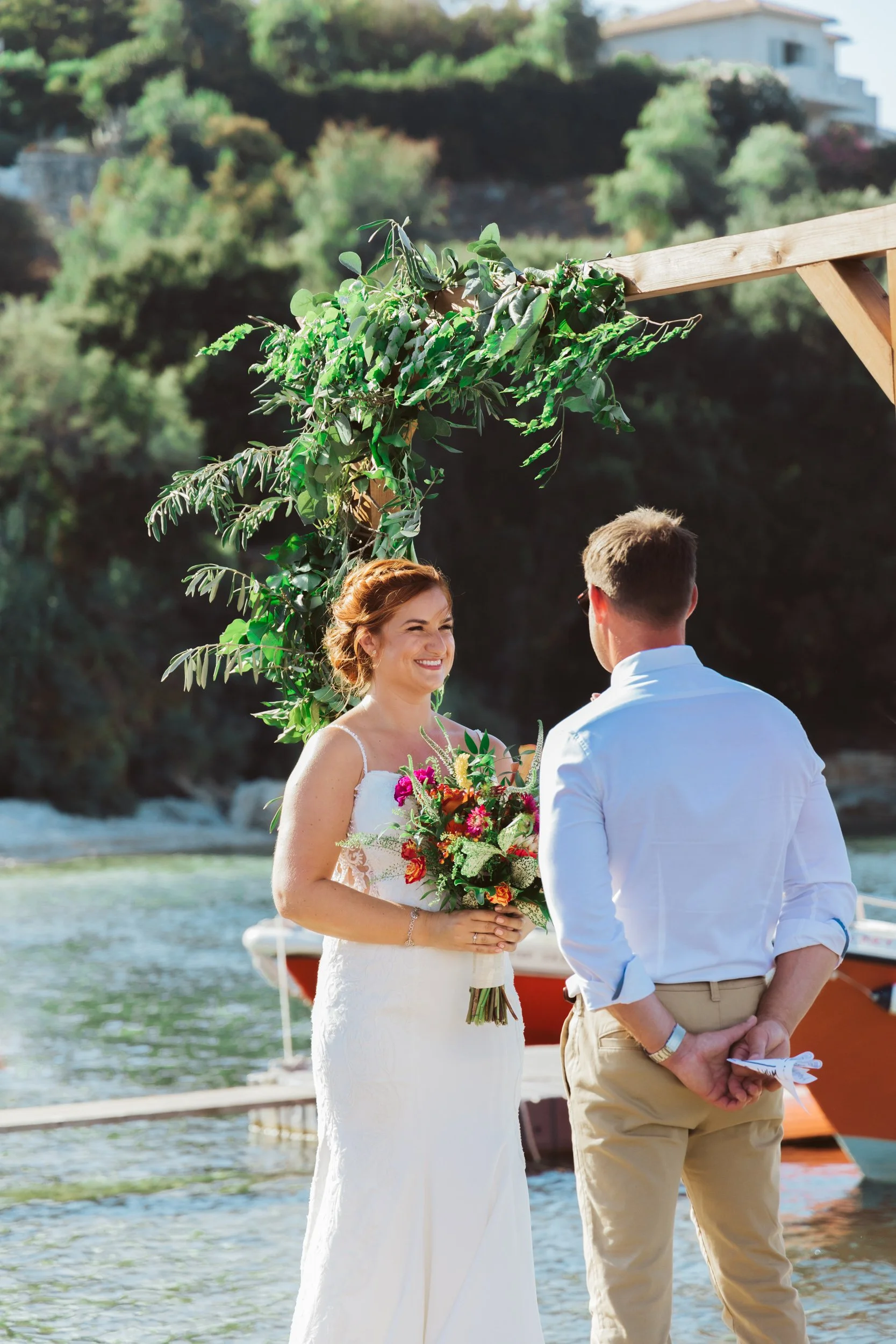A bride and groom exchanging vows outdoors by the water, with a floral arch and boats in the background.