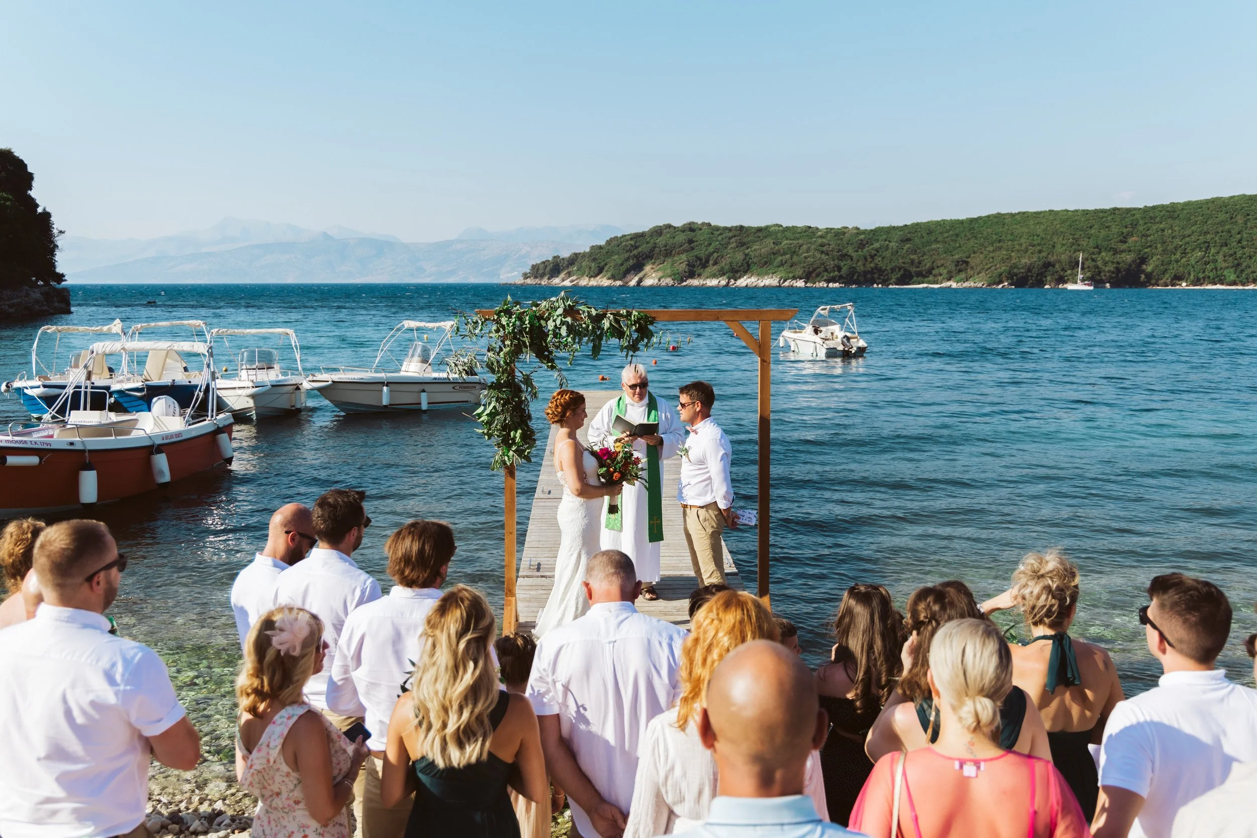A wedding ceremony taking place on a dock by the water, with a bride and groom standing under a wooden arch decorated with greenery, officiated by a priest. Guests are seated and standing along the rocky shoreline, with boats anchored nearby in a sce
