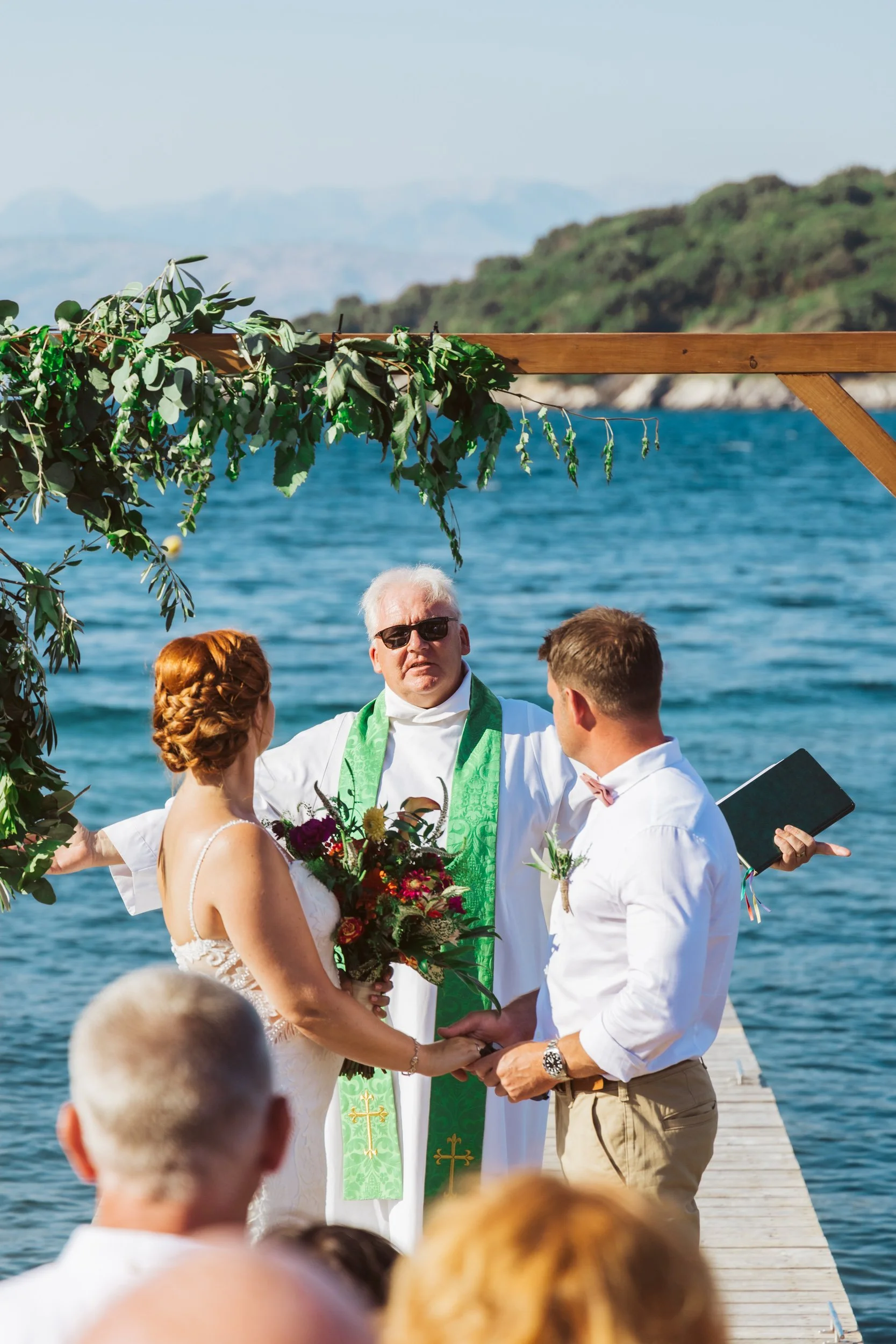 A wedding ceremony taking place outdoors on a dock near water, with a priest officiating as a bride and groom exchange vows surrounded by guests, and a floral arrangement above.