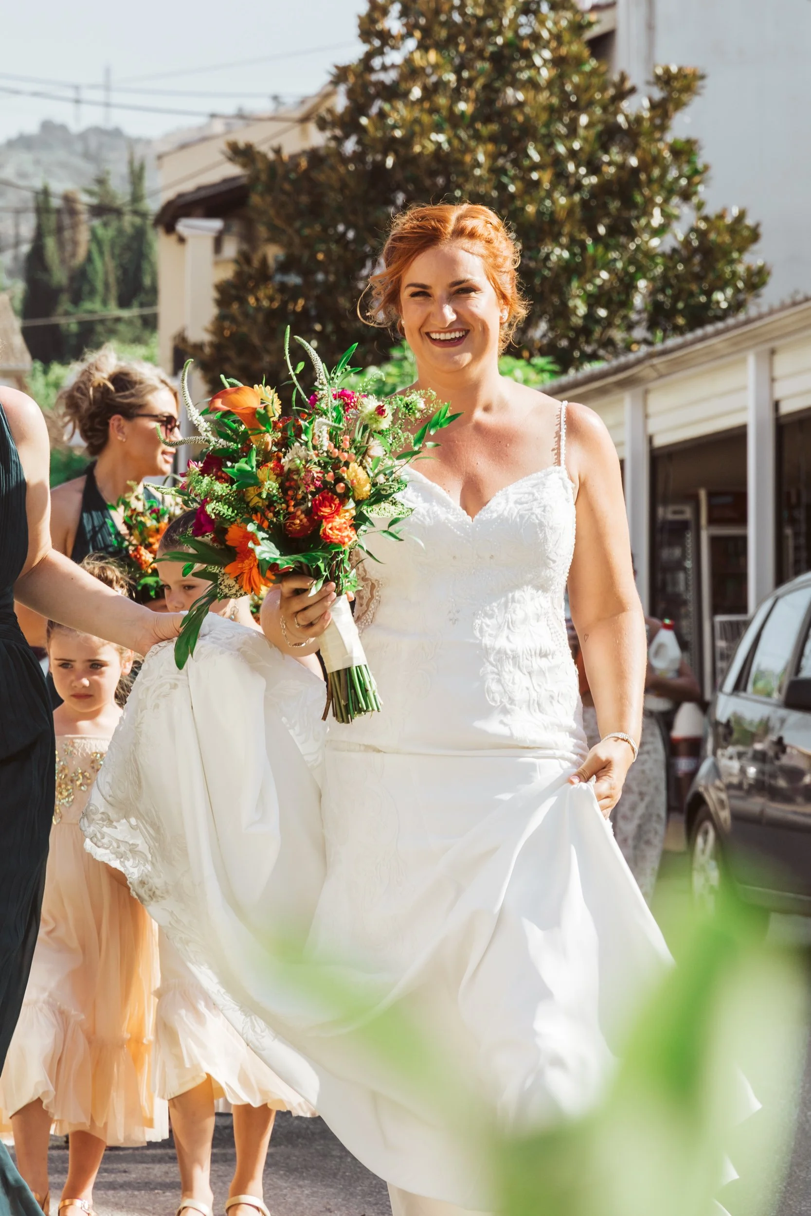A smiling bride in a white wedding dress holding a colorful bouquet walking outdoors on a sunny day.