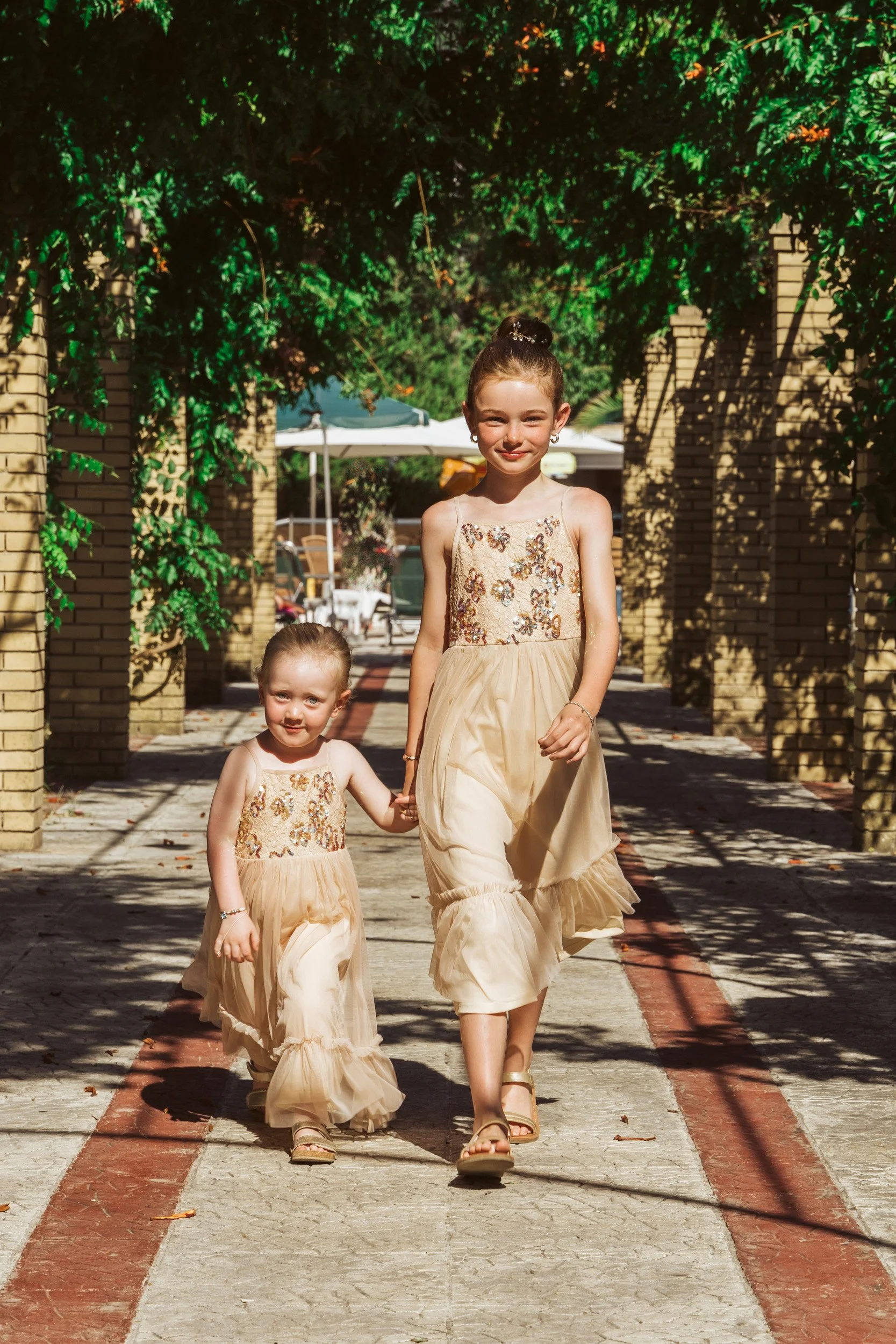 Two young girls wearing matching gold dresses walking hand in hand outdoors on a sunny day, with greenery overhead and brick pillars on either side.