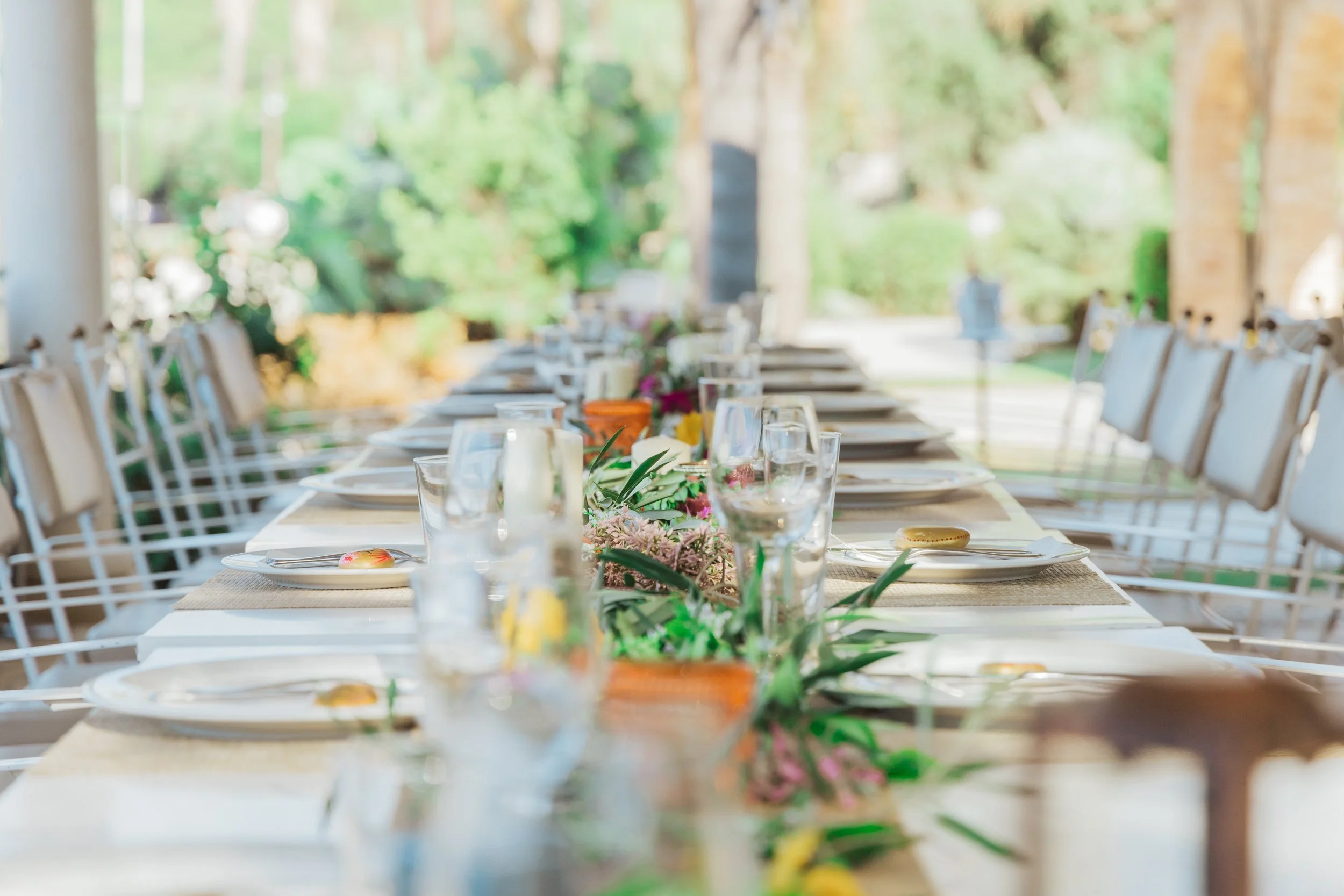 A long outdoor dining table set with plates, glasses, cutlery, and floral centerpieces on a sunny day.