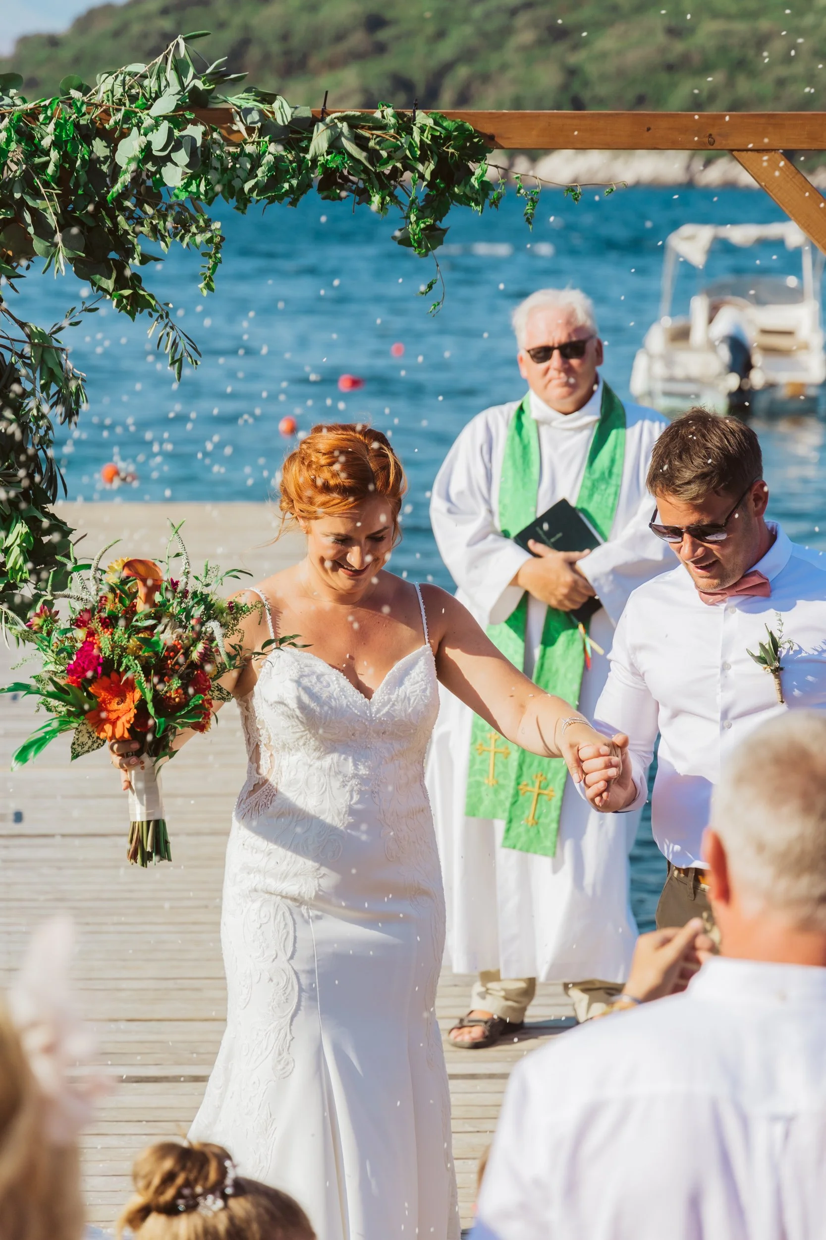 A bride and groom exchanging vows during their wedding ceremony on a dock near a lake, surrounded by guests. The bride is holding a bouquet of colorful flowers, and the officiant stands behind them.