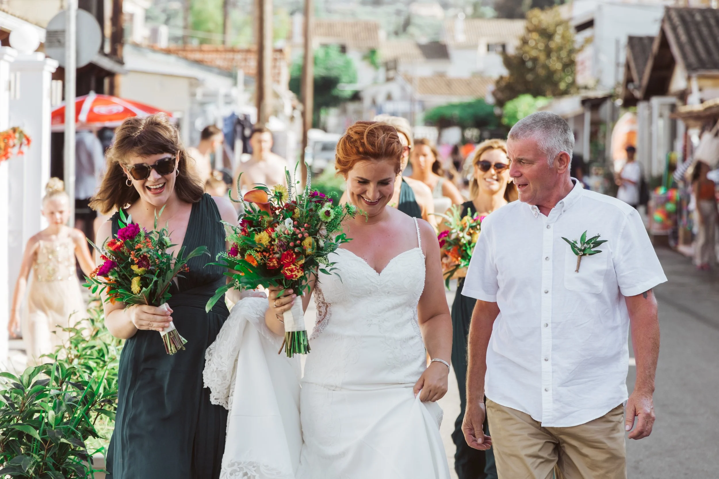 A bride in a white wedding dress holding a bouquet of flowers, walking and smiling with her father and bridesmaids on a lively street during a wedding celebration.