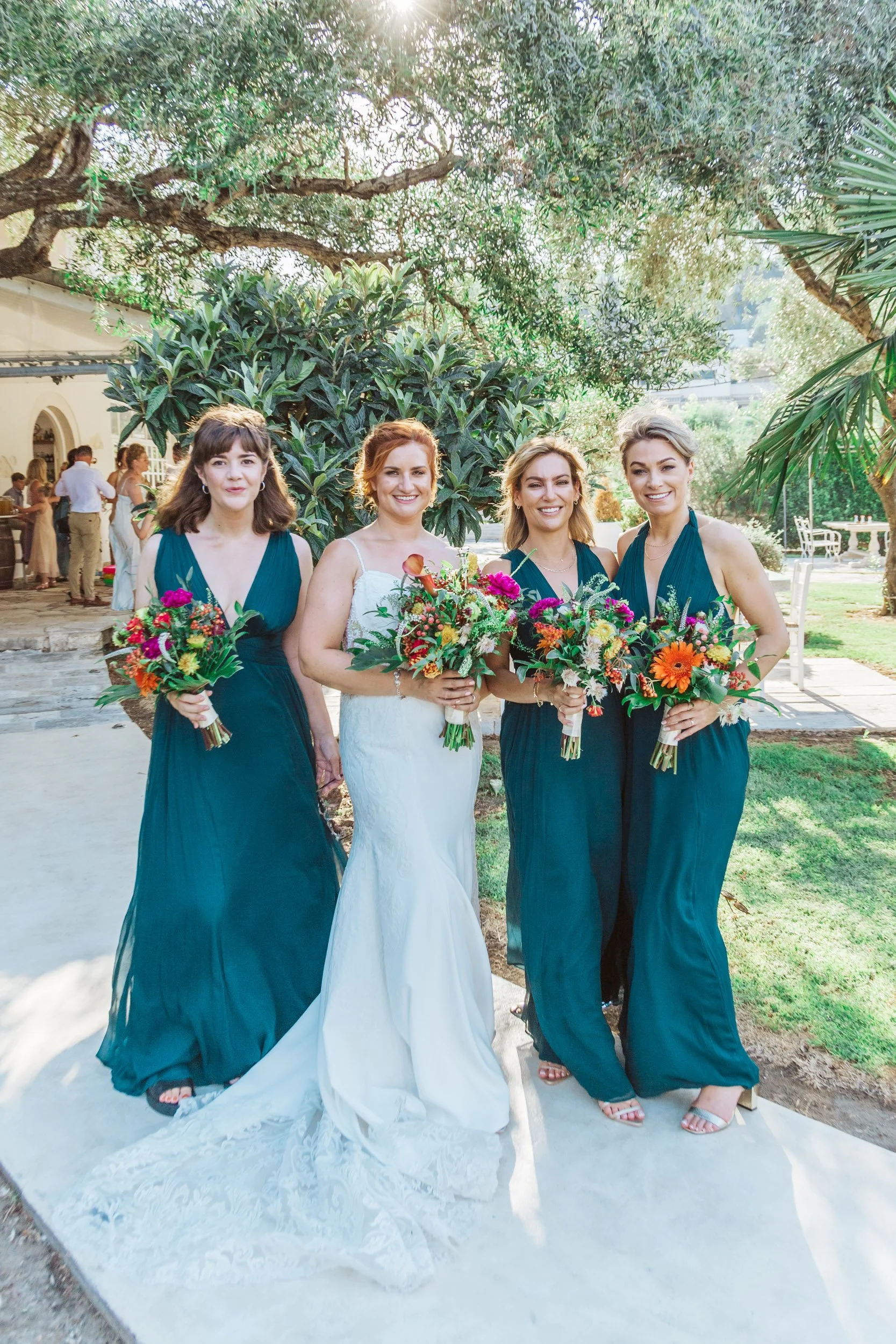Four women at a wedding, three bridesmaids in matching teal dresses and a bride in a white dress, all holding colorful bouquets, outdoors with trees and greenery.